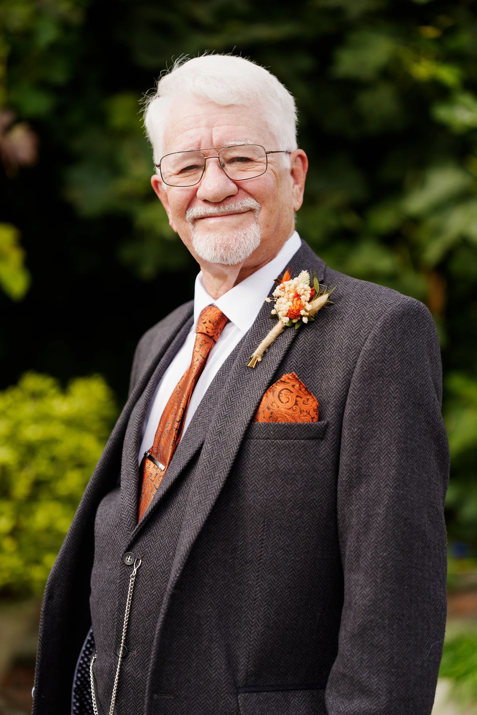 A smiling older gentleman with white hair and glasses poses outdoors wearing a charcoal three-piece herringbone suit, an orange patterned tie, a pocket square, and a coordinating boutonniere.