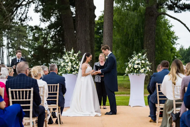 The bride and groom exchange rings during an outdoor ceremony set against a backdrop of tall trees and white floral pedestal arrangements.