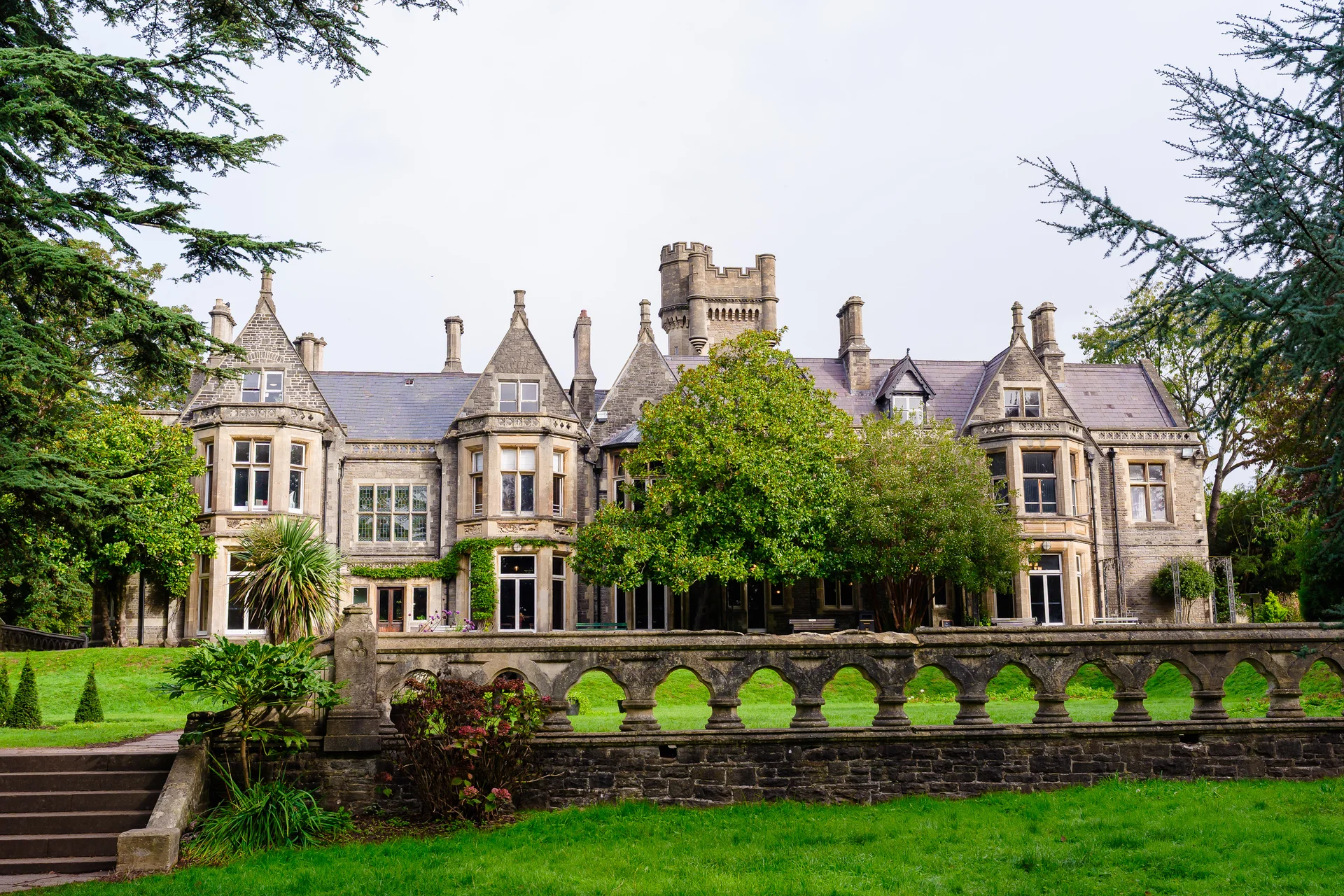 A grand historic stone mansion and castle venue stands amidst lush green lawns and mature trees under an overcast sky.