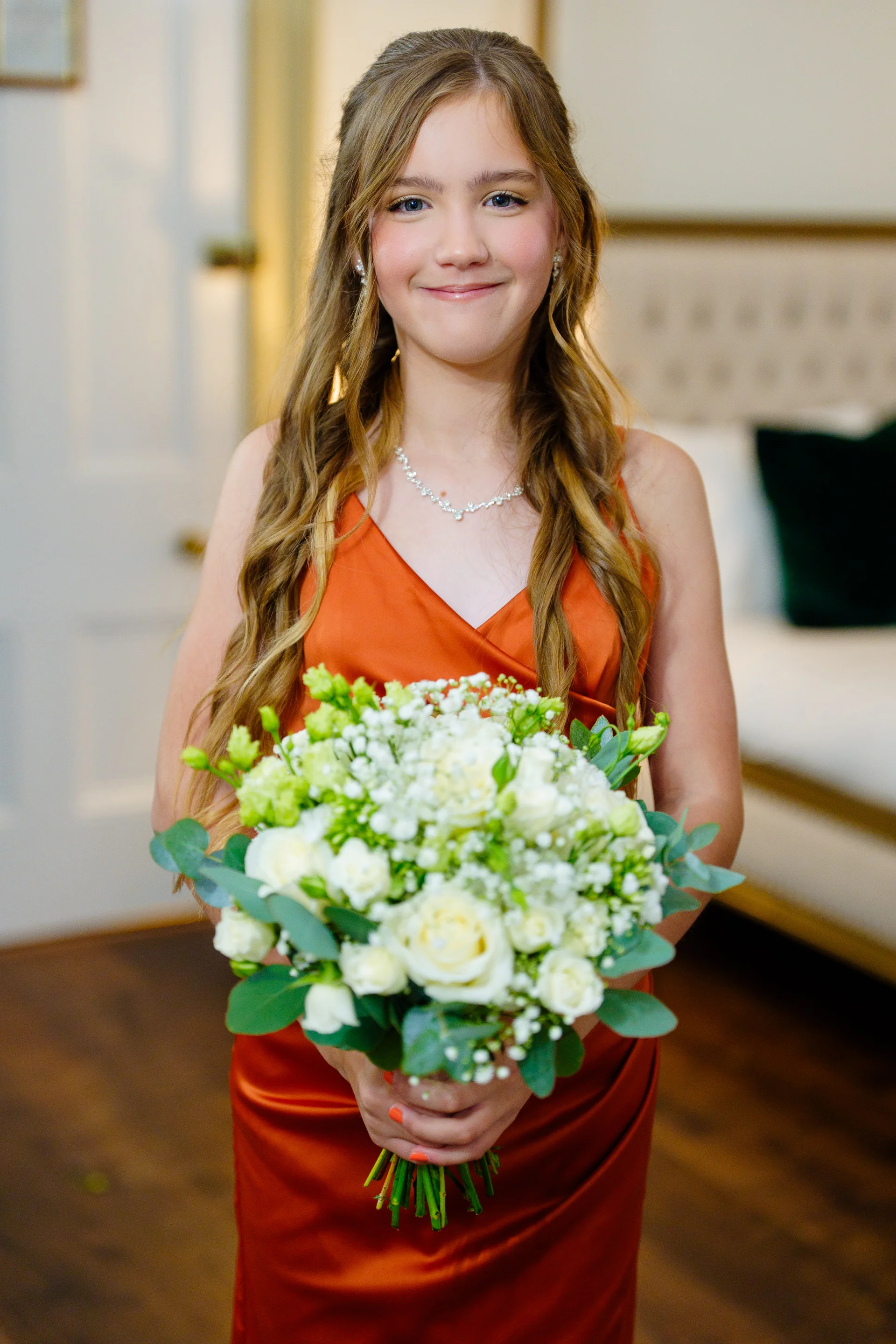 A young bridesmaid wearing a rust-coloured dress and holding a hand-tied bouquet of white roses, baby's breath, and eucalyptus.