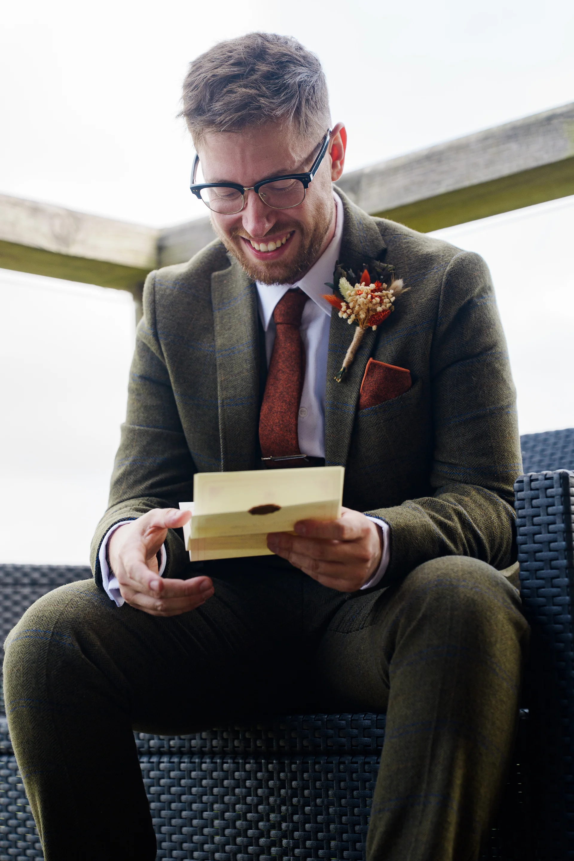 A smiling groom wearing a green tweed suit and glasses sits outside while reading a heartfelt letter before his wedding.