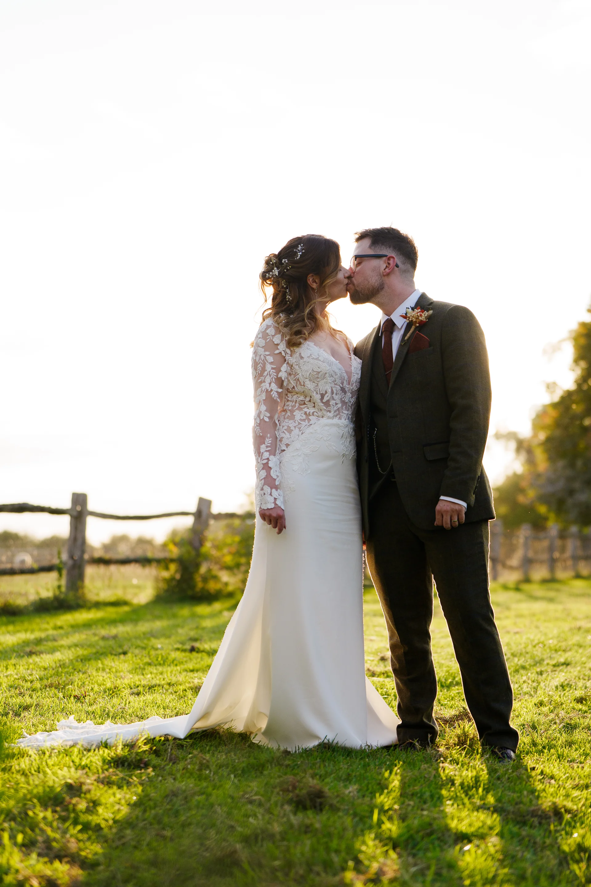 A newlywed couple shares a tender kiss bathed in warm golden hour sunlight during an outdoor portrait session in a scenic countryside field.