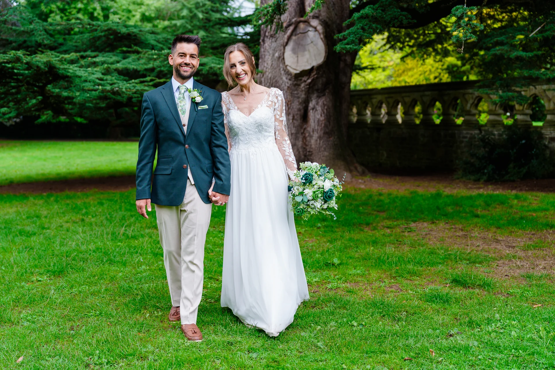 A newlywed couple happily walk hand in hand across a lush green lawn in a garden setting, with the bride holding a white and green bouquet and wearing a long-sleeved lace wedding dress, while the groom wears a dark green jacket and light trousers.