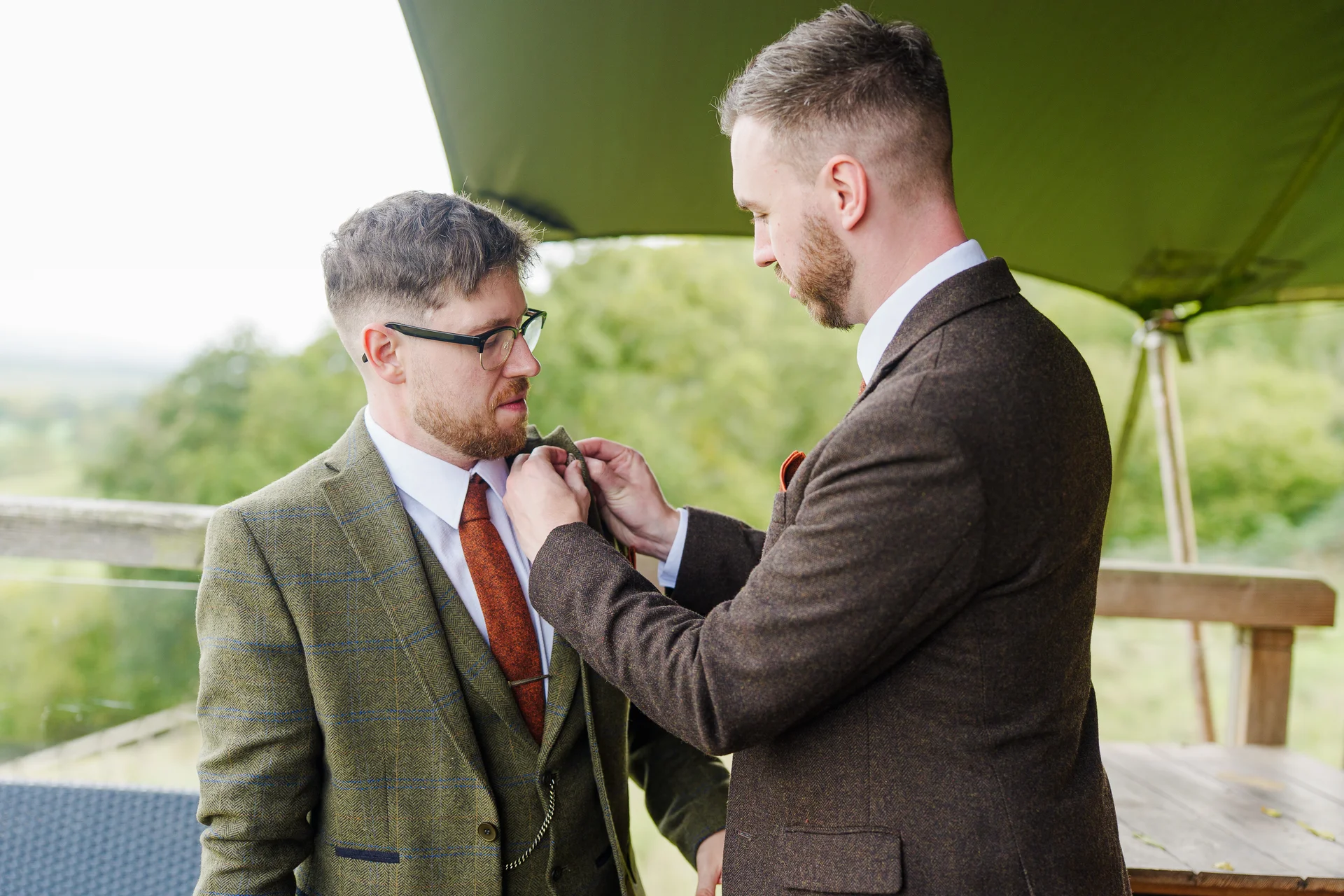A groom helping another man adjust his tweed suit jacket outdoors on their wedding day.