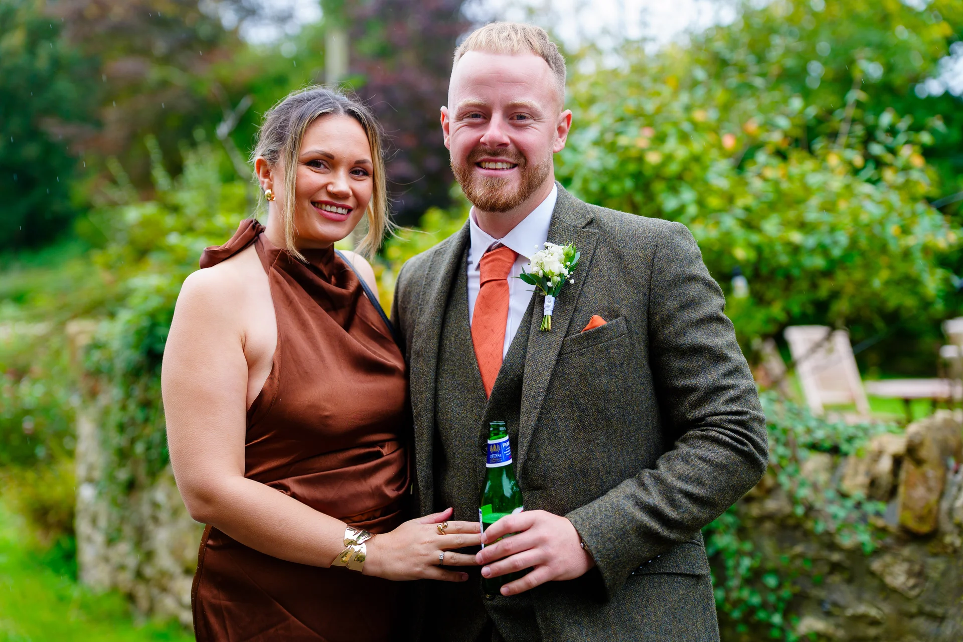 A smiling wedding guest in a satin brown dress and a groom in a tweed suit stand outdoors holding a beer bottle.