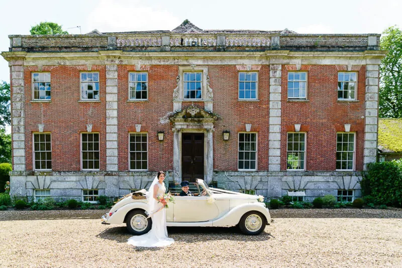 A smiling bride holding a bouquet stands beside a classic cream vintage wedding car parked on a gravel driveway in front of a grand Georgian manor house.