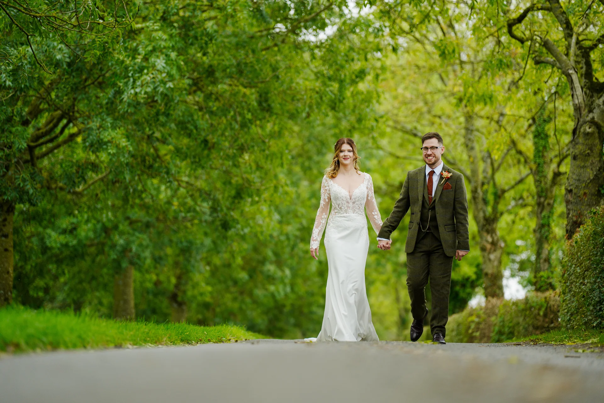 A newlywed couple holds hands and smiles as they walk along a country path lined with lush green trees.