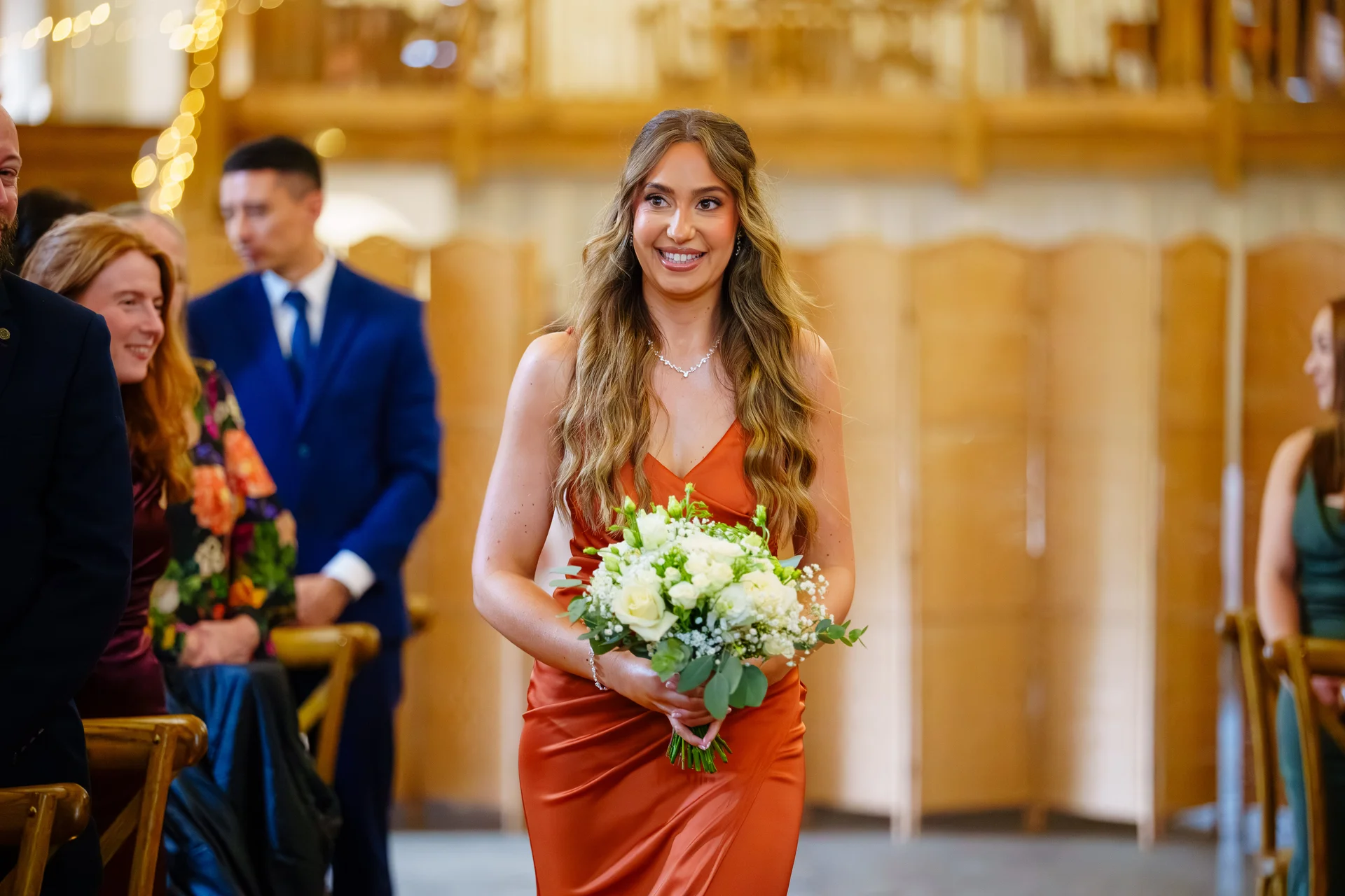 A smiling bridesmaid in a rust-coloured satin dress walks down the aisle carrying a hand-tied bouquet of white roses and eucalyptus.