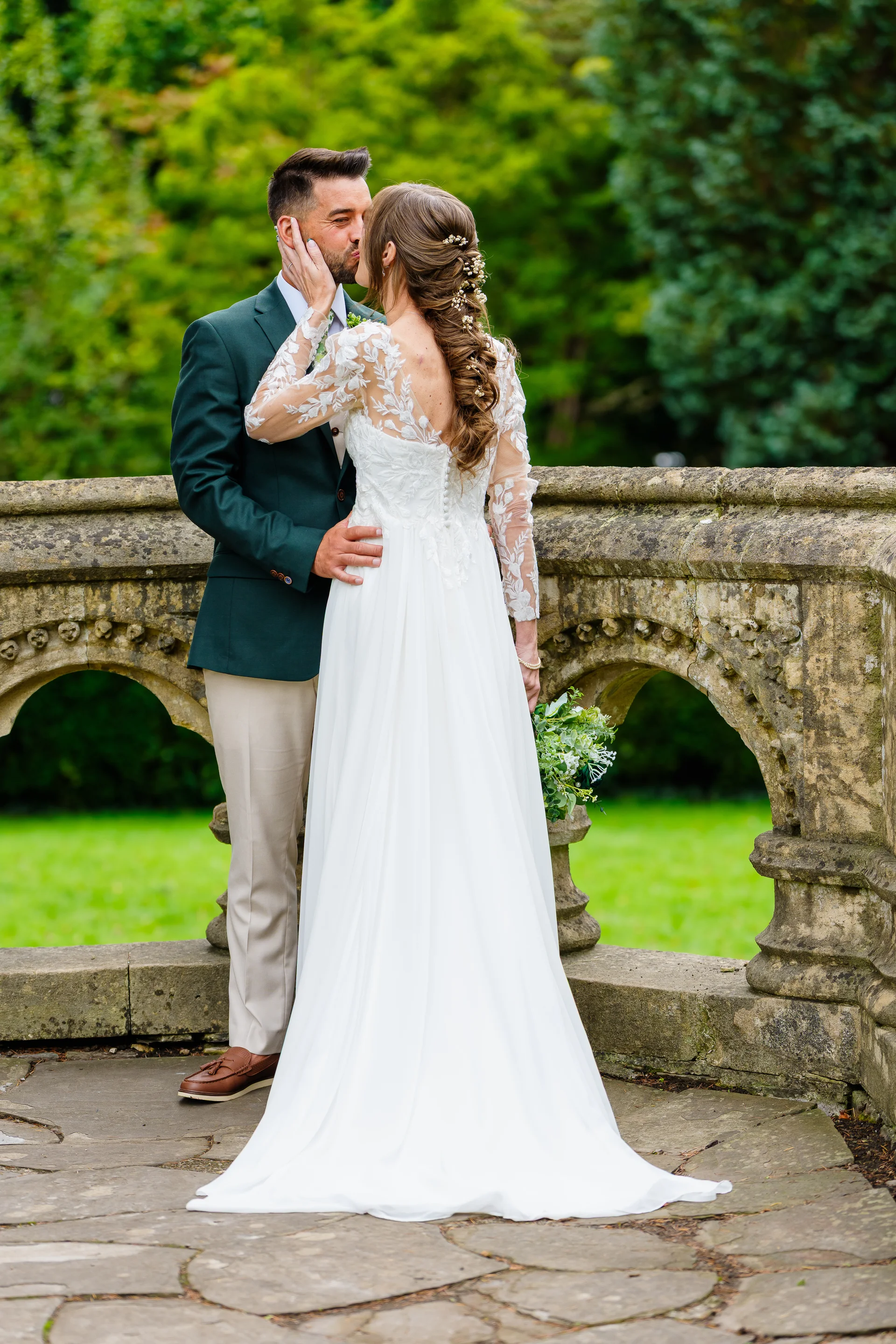 Newlyweds share a tender kiss on a stone bridge framed by lush green foliage during their outdoor wedding celebration.