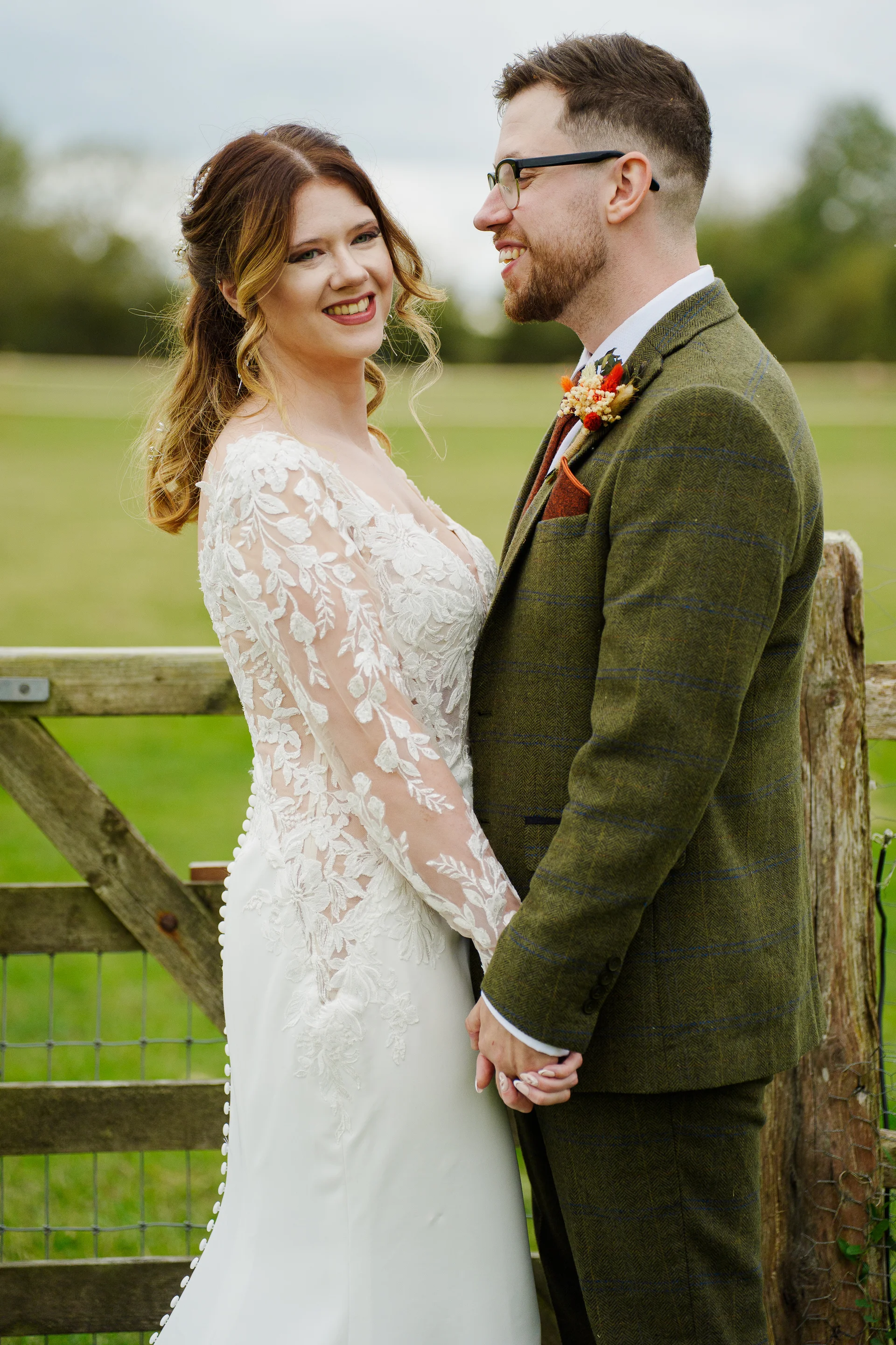 A smiling bride and groom holding hands next to a wooden farm gate in a lush green field.