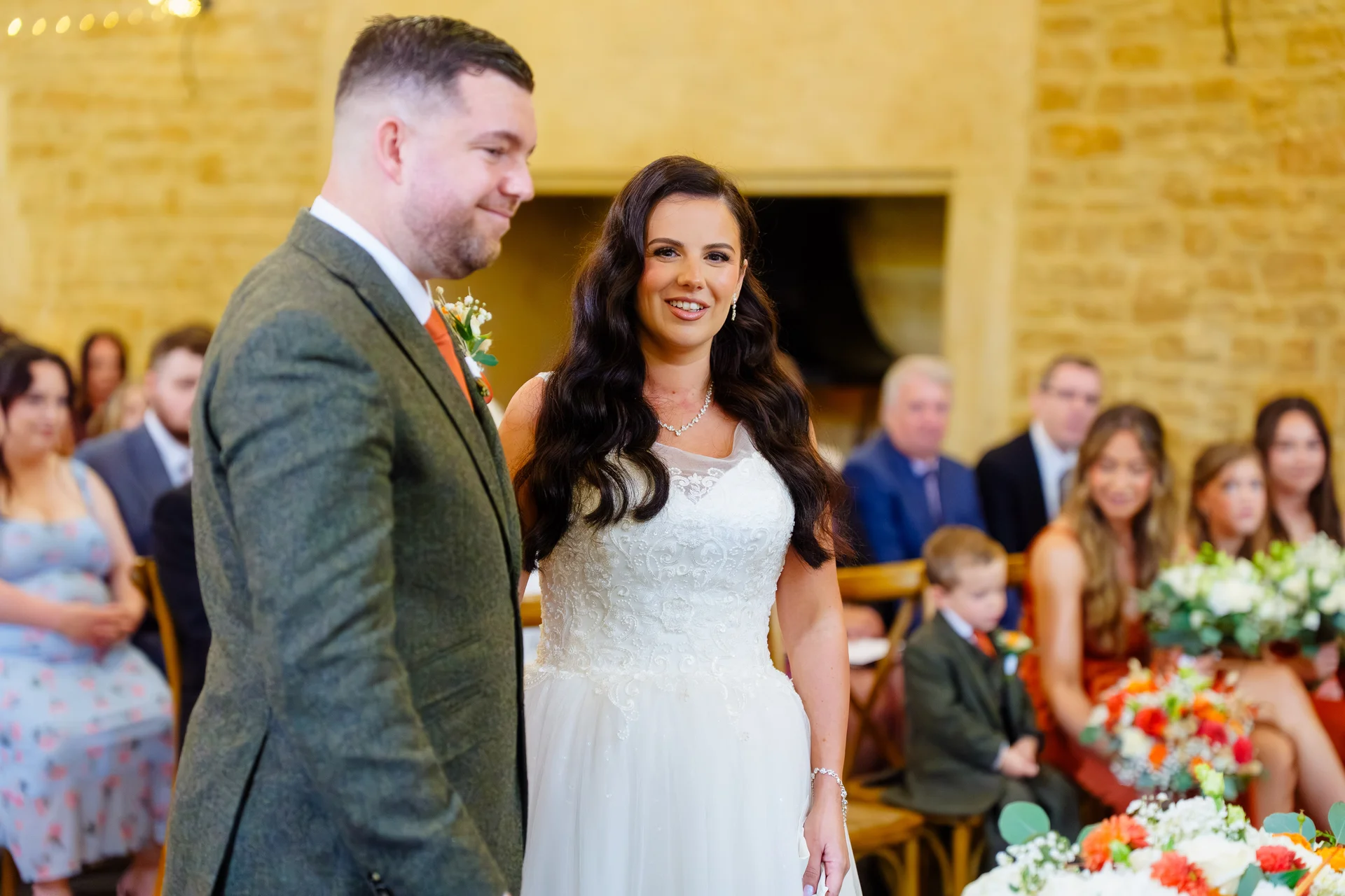 A smiling bride and groom stand together at the front of a rustic stone barn wedding ceremony, surrounded by seated guests and floral arrangements.