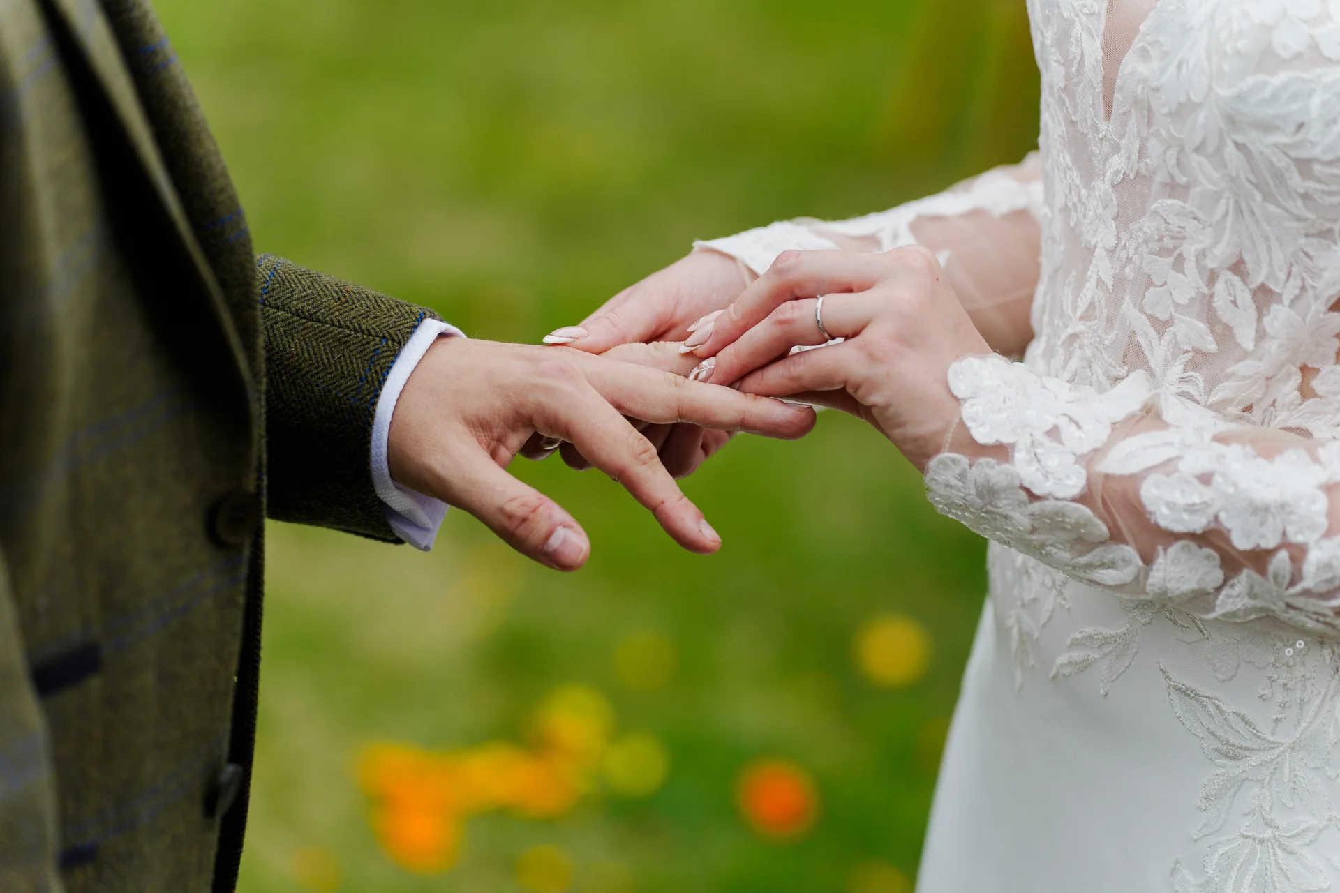 A close-up shot captures the bride placing a wedding ring onto the groom's finger outdoors, showcasing her lace sleeve and his tweed suit jacket.