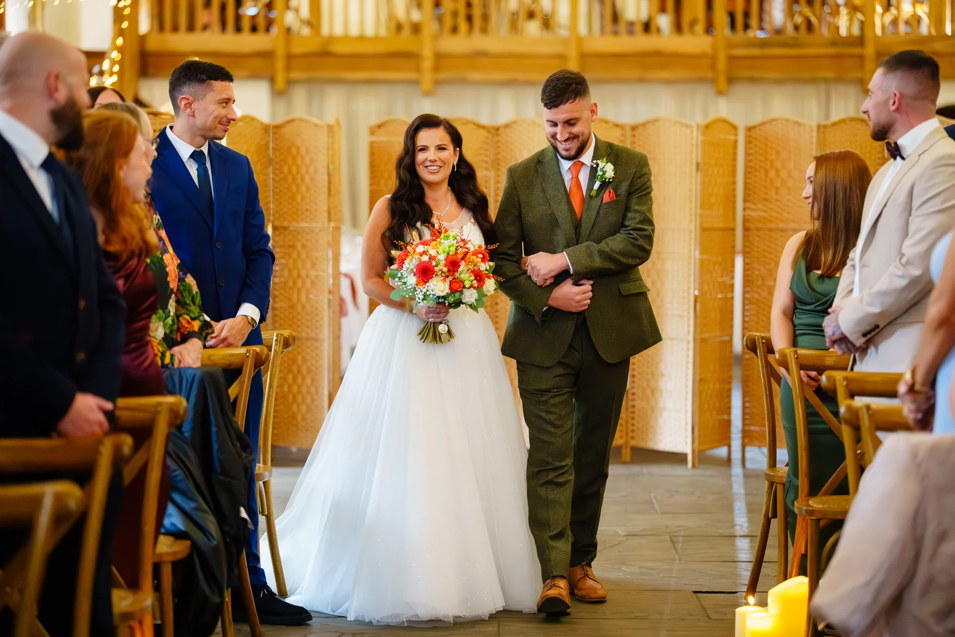 A smiling bride and groom walk hand in hand down the aisle of a rustic wooden barn venue, surrounded by seated wedding guests.