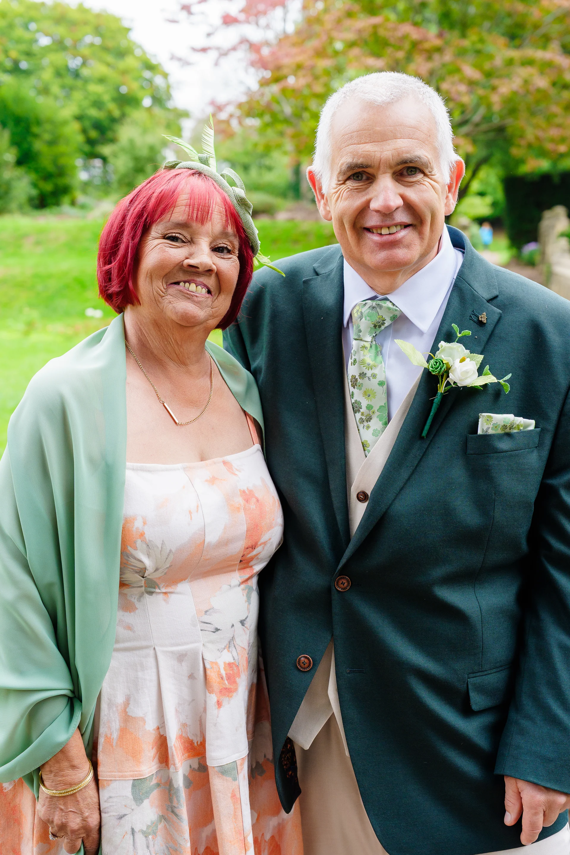 An older couple smiles warmly for a posed portrait outdoors in a lush green garden during a wedding celebration.