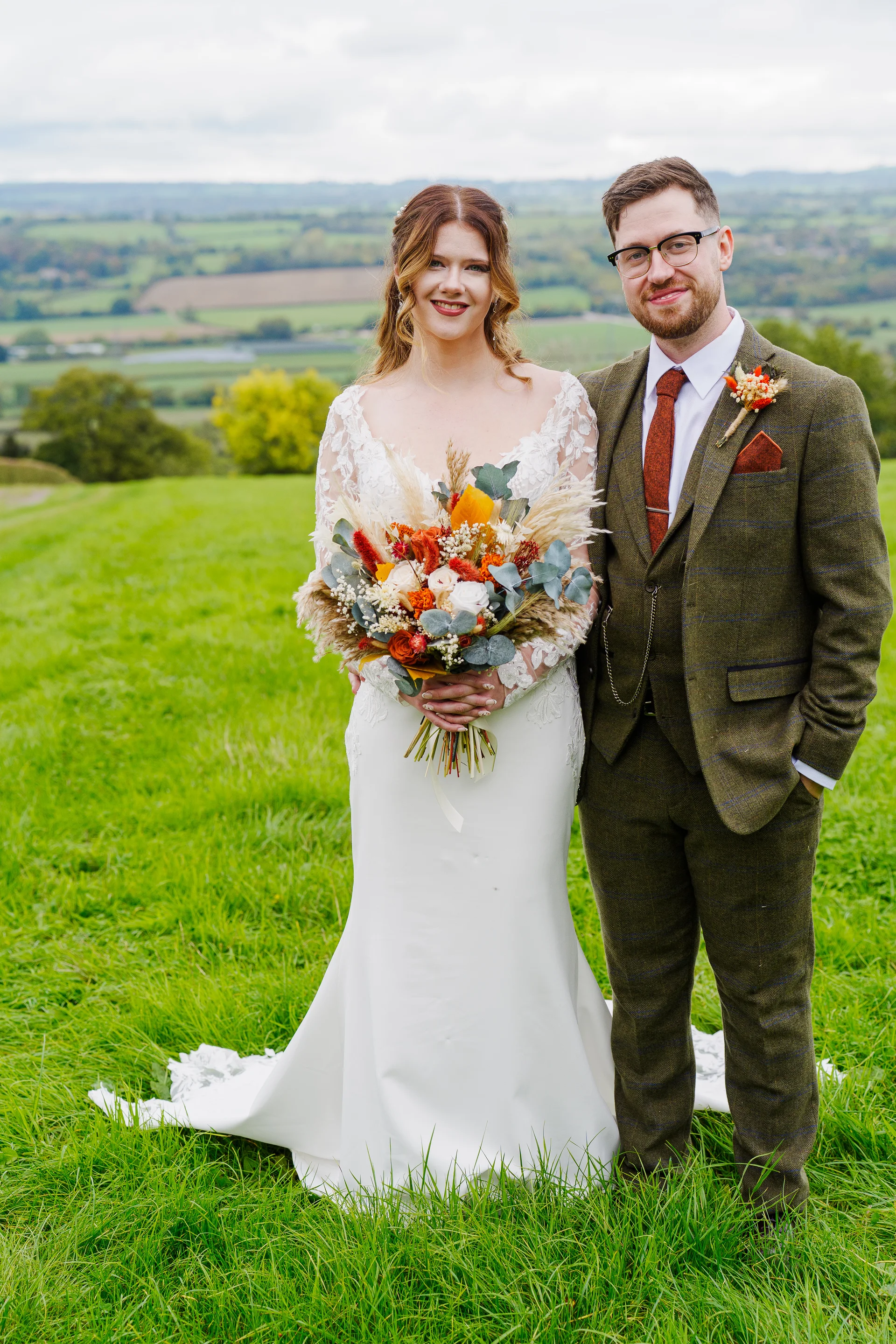 A smiling bride and groom pose in a lush green field with rolling hills in the background, holding an autumnal bouquet containing burnt orange flowers and pampas grass.