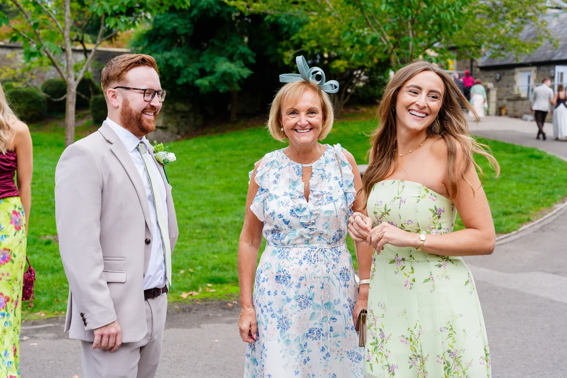 A smiling group of wedding guests enjoying a chat outdoors near a green lawn, featuring a woman in a floral dress and a man in a beige suit.