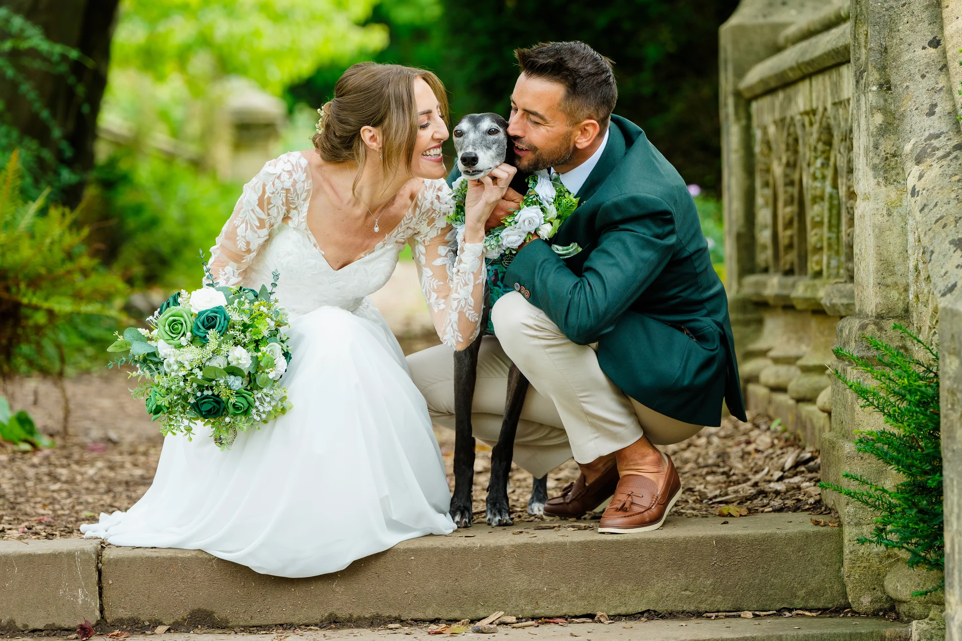 A joyful bride and groom laugh as they cuddle their greyhound wearing a floral collar on stone steps outdoors.