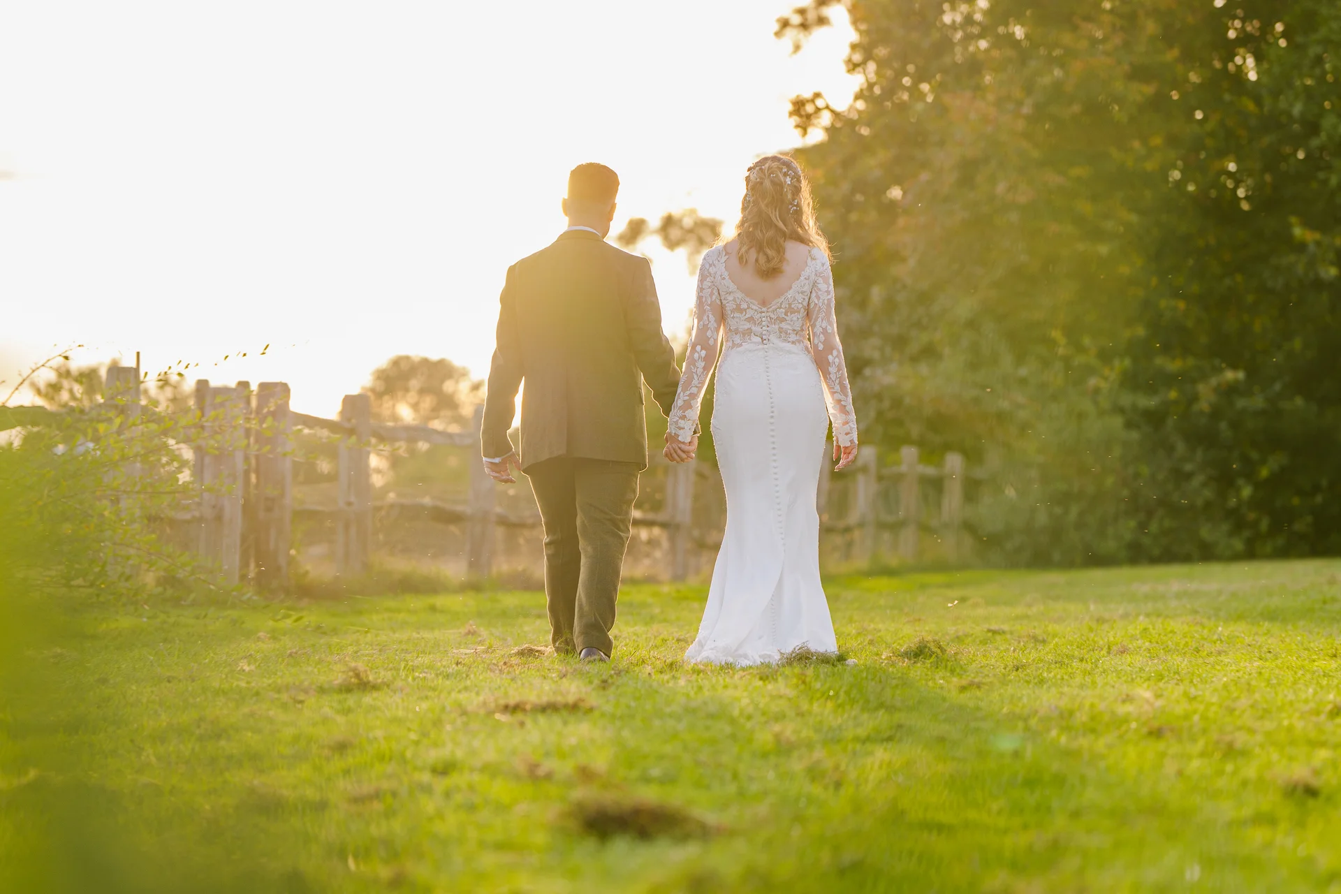 A newlywed couple walking hand in hand through a grassy field at sunset, illuminated by warm golden light against a rustic wooden fence.