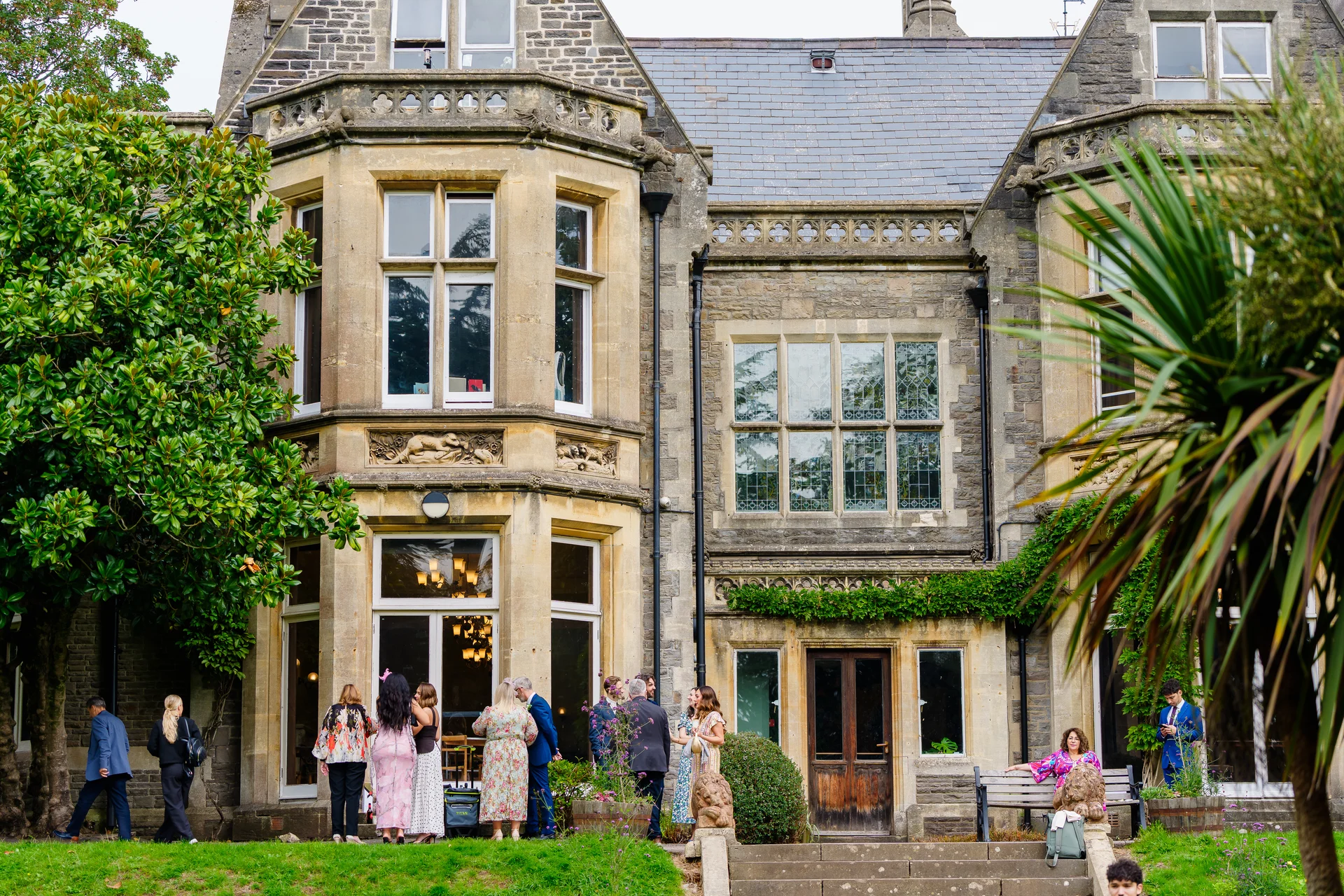 Wedding guests mingle outside on the lawn in front of a grand historic stone country house during the drinks reception.