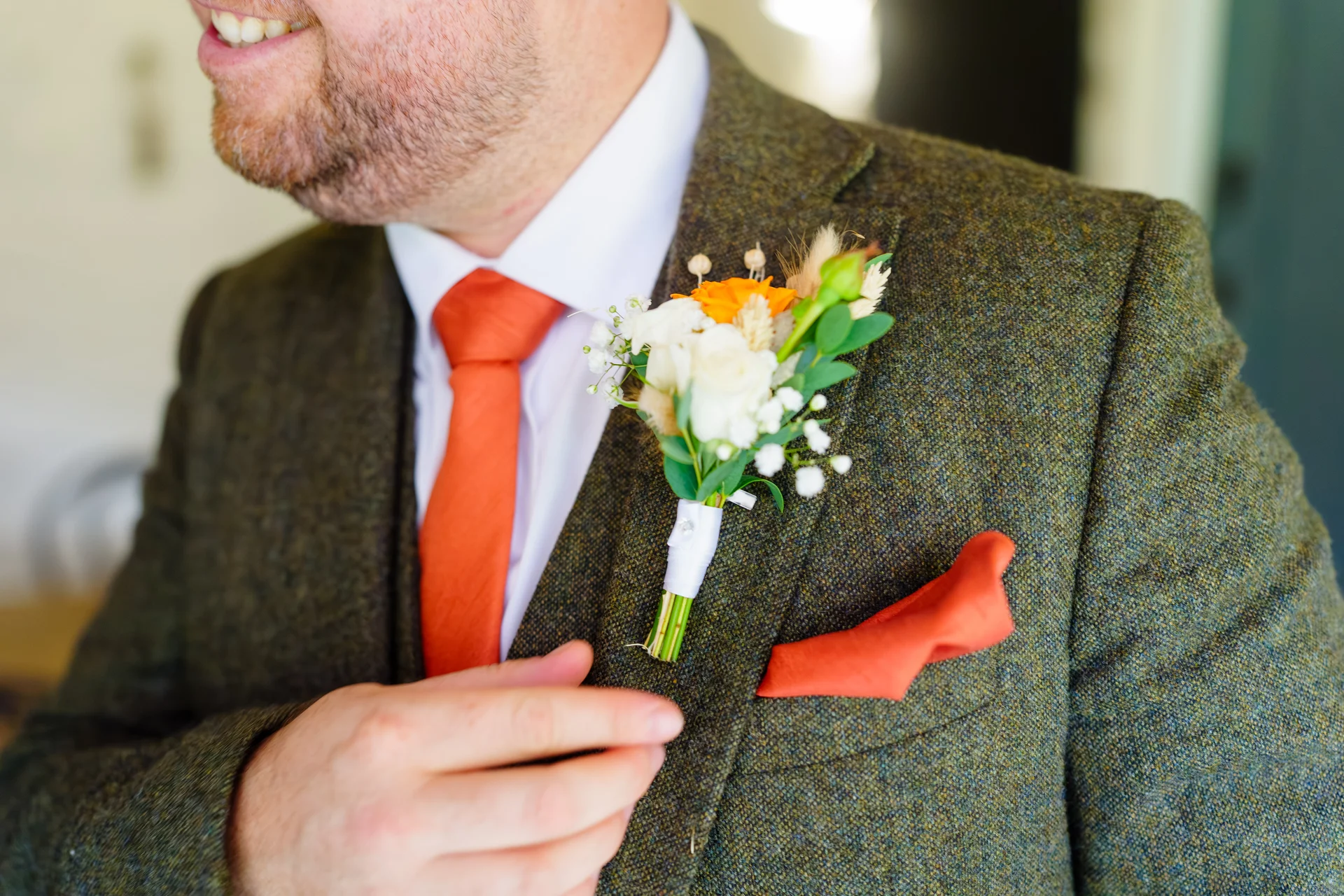 Close-up portrait of a groom wearing a green tweed suit and waistcoat, accented with a vibrant burnt orange tie, pocket square, and a coordinating floral buttonhole.
