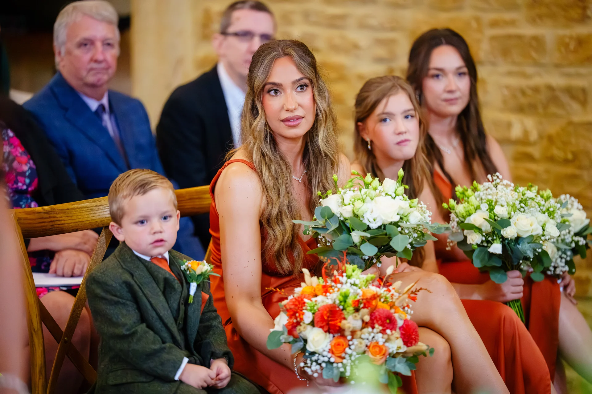 A wedding party seated together on wooden chairs, featuring bridesmaids in rust orange dresses holding floral bouquets alongside a young page boy in a green tweed suit.