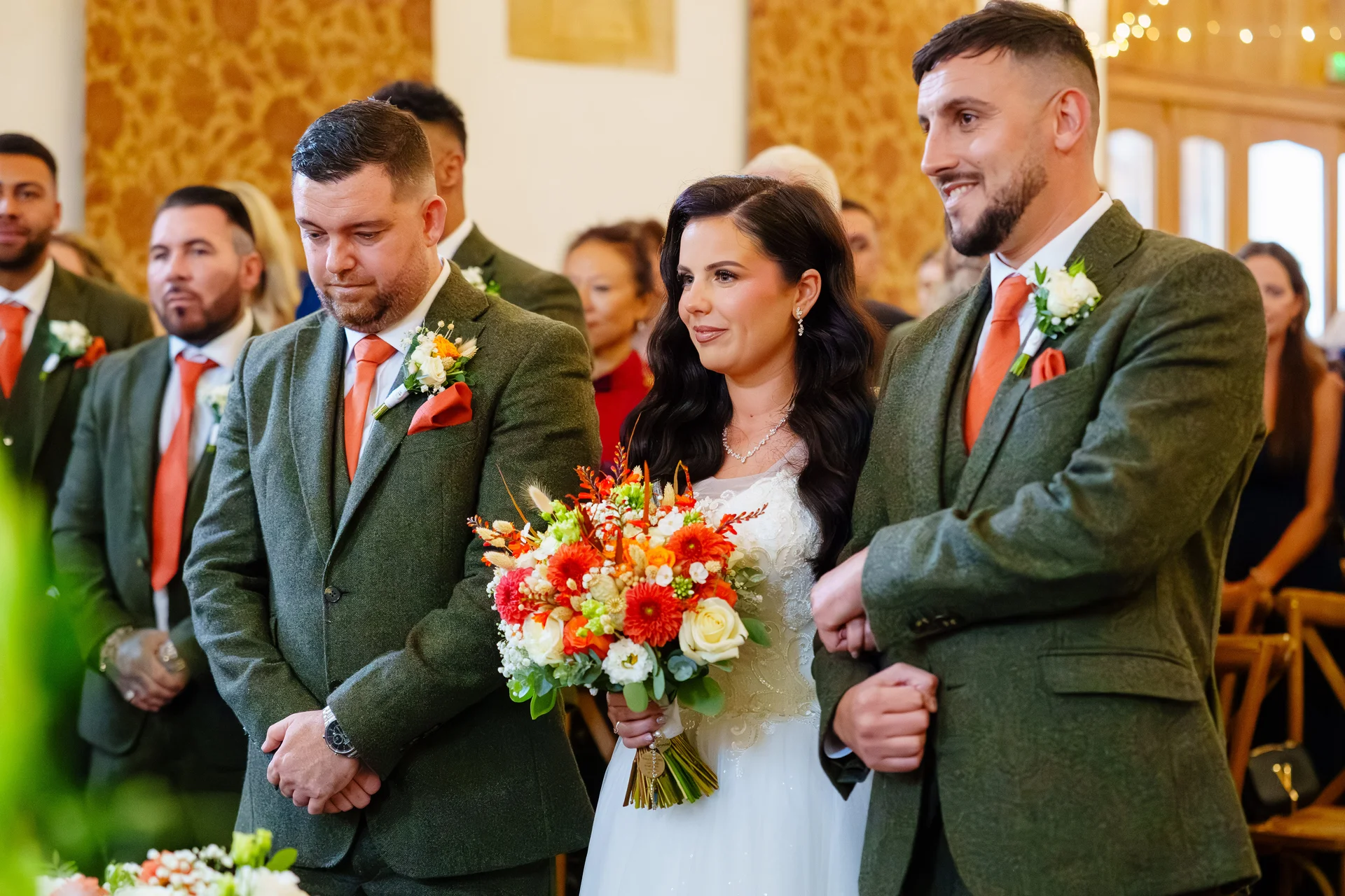 A smiling bride holding a vibrant orange and white bouquet stands between the groom and best man, both dressed in sharp green tweed suits with matching orange ties during the wedding ceremony.