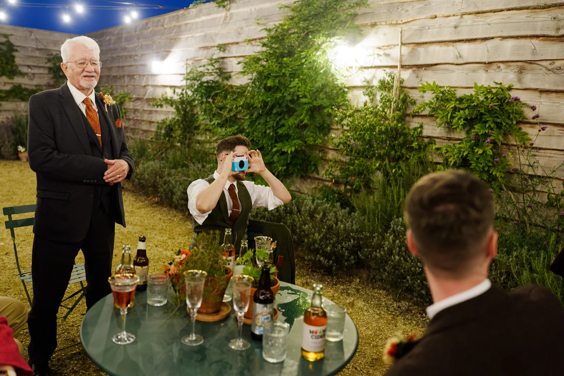 An older man in a suit looks smilingly toward a seated guest who is playfully framing a photo with a small blue camera across a garden table during an evening wedding reception.