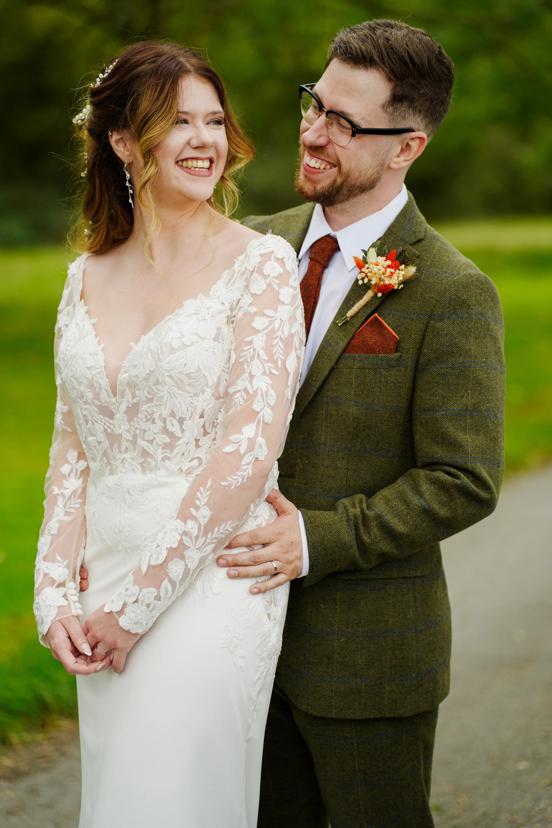 A smiling bride in a lace-sleeved gown stands beside her groom, who wears a green tweed suit and a boutonniere, outdoors during their wedding portrait session.