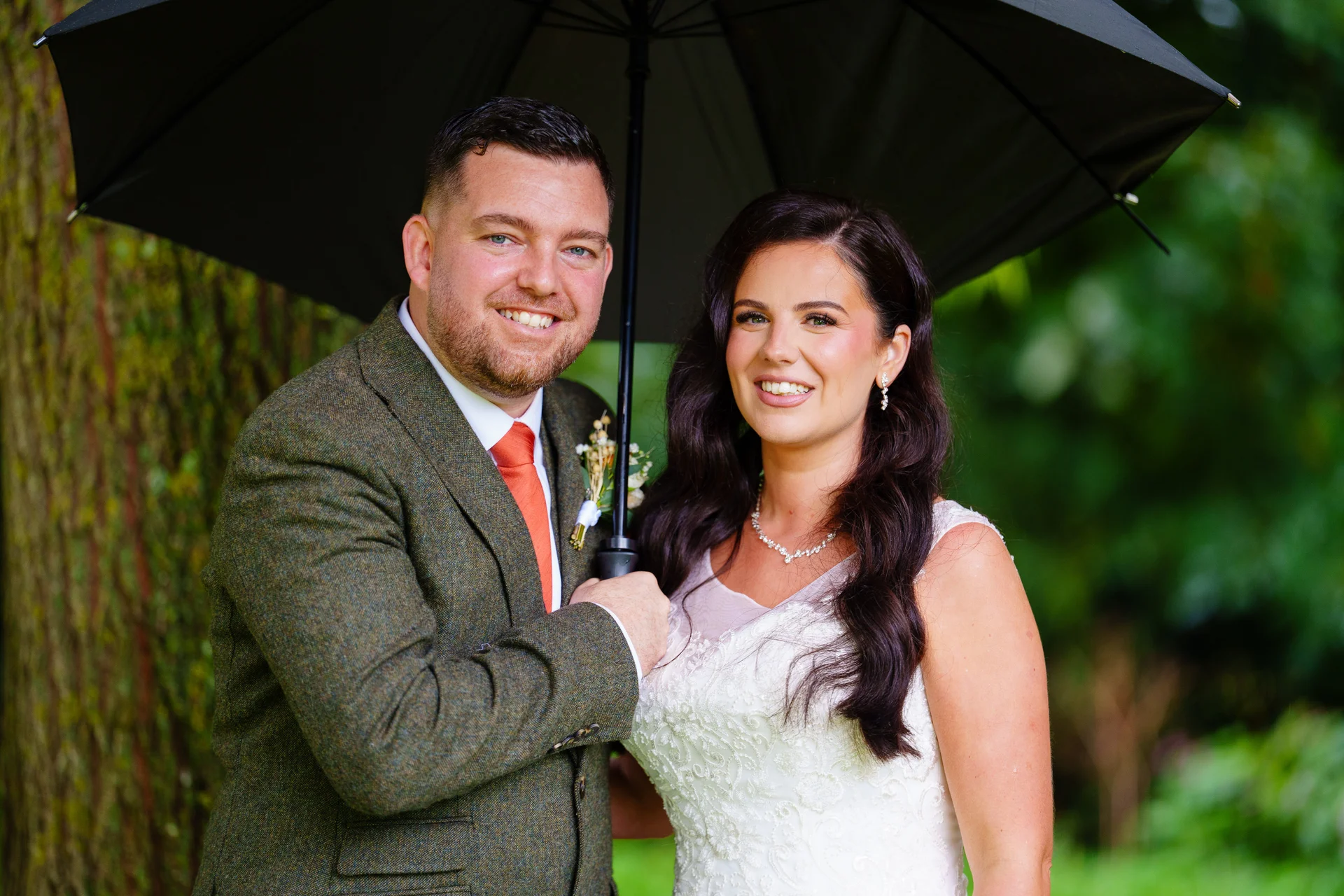 A smiling bride and groom pose together under a black umbrella outdoors on their rainy wedding day.