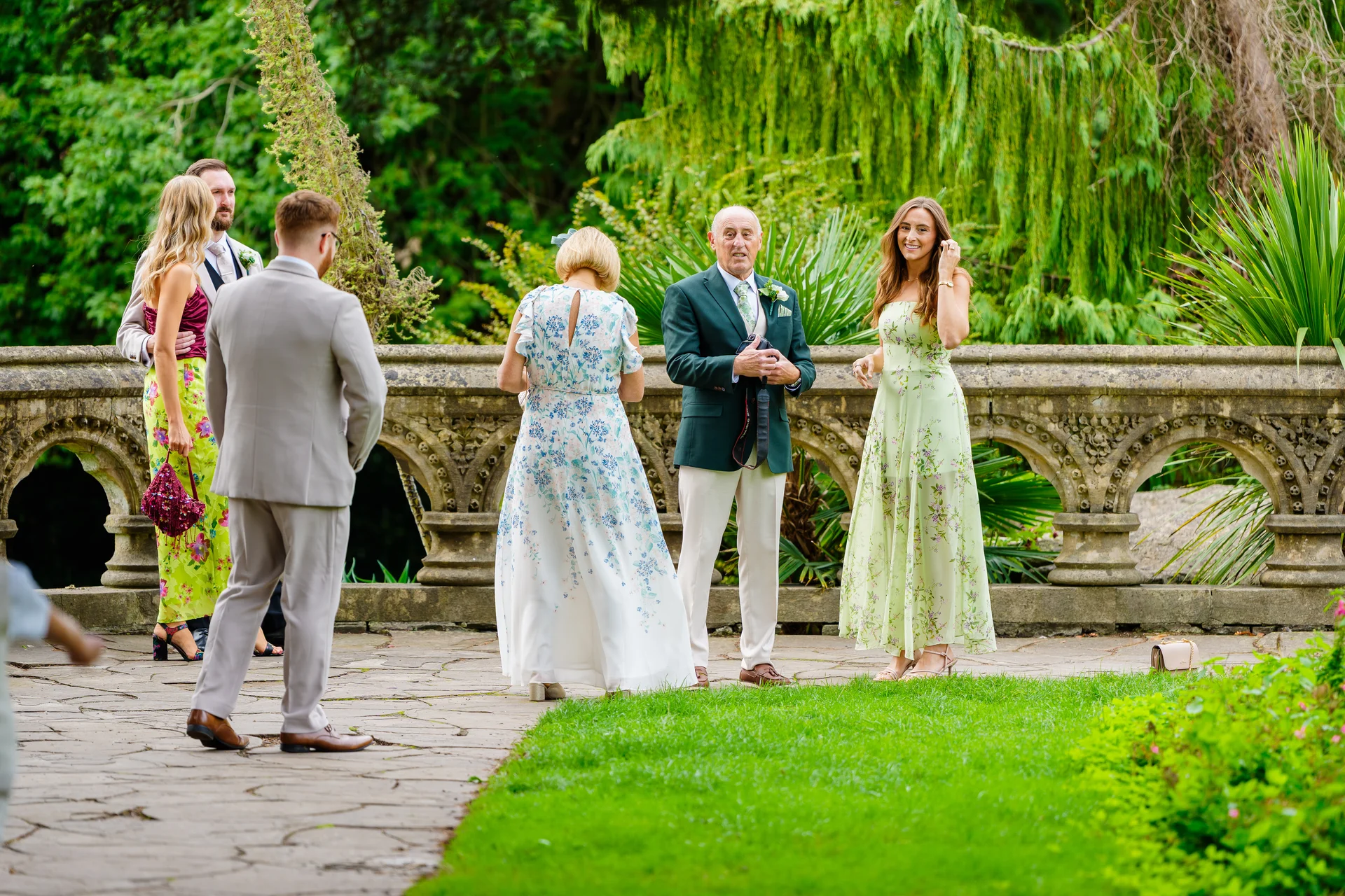 Wedding guests mingle outdoors in a lush garden setting by an ornate stone balustrade, dressed in colourful attire and suits.