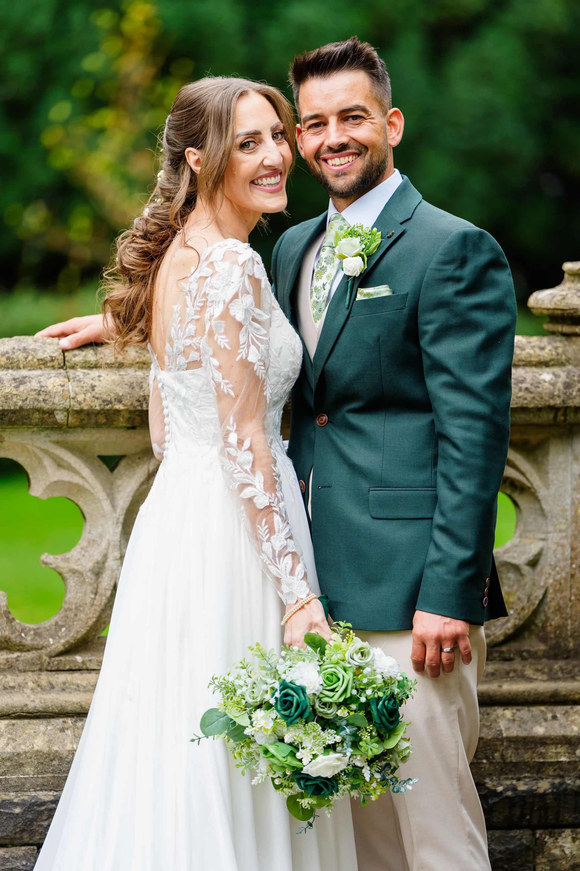 A smiling bride holding a green and white bouquet stands alongside her groom in a dark green suit against an outdoor stone bridge backdrop.
