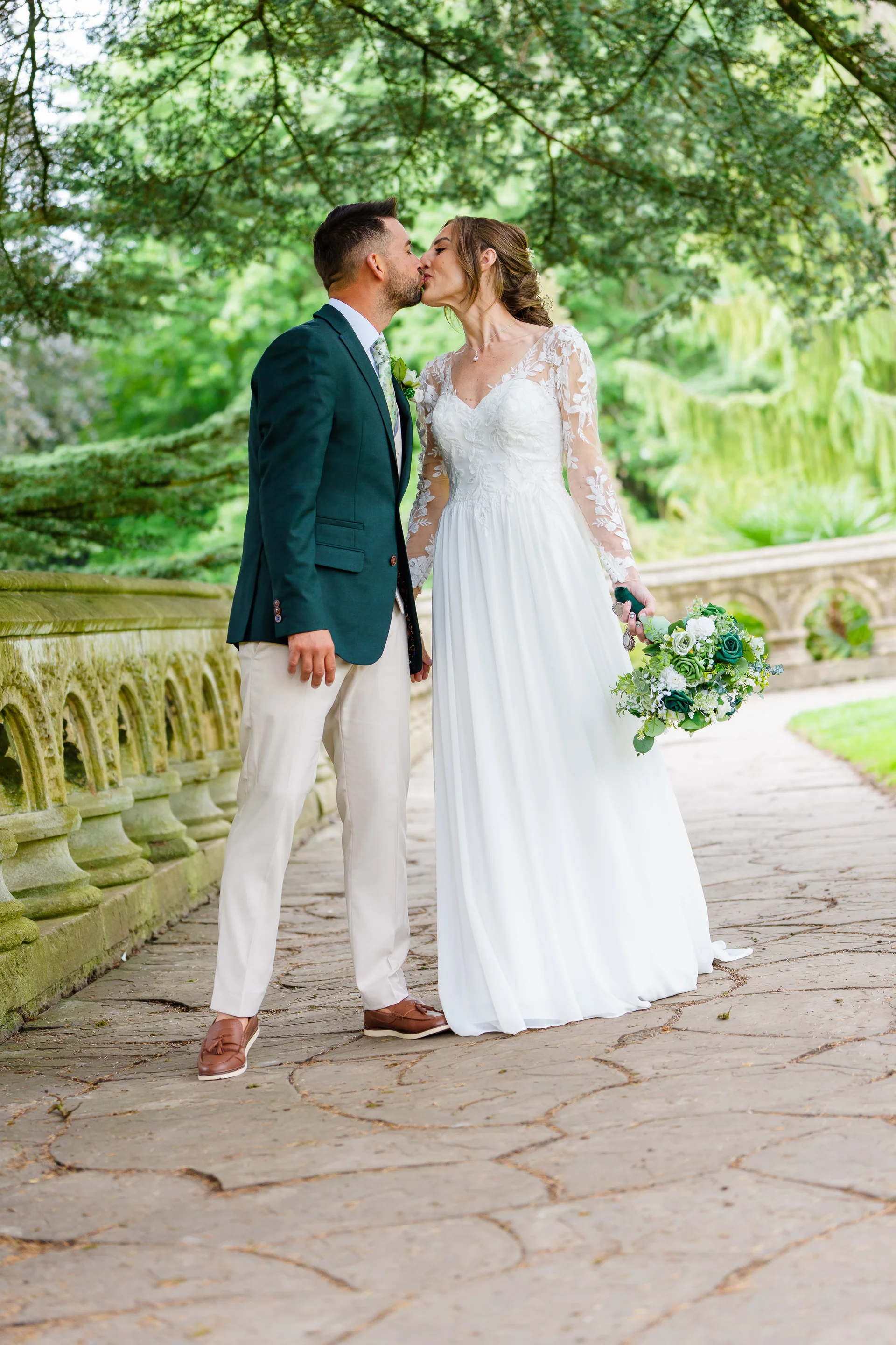 A newlywed couple shares a tender kiss on a stone pathway framed by lush green trees and an ornate balustrade.
