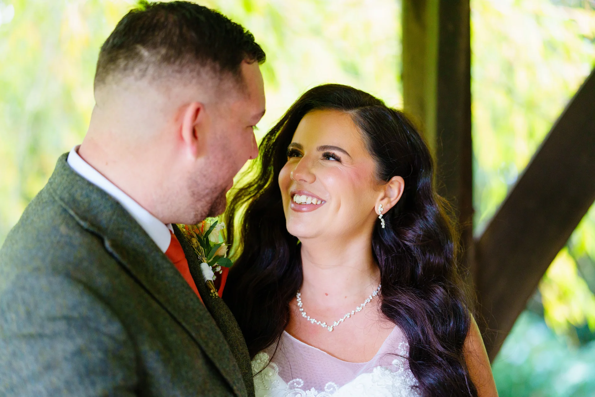 A smiling bride with long dark wavy hair looks lovingly at her groom in a tweed suit during an outdoor portrait session.