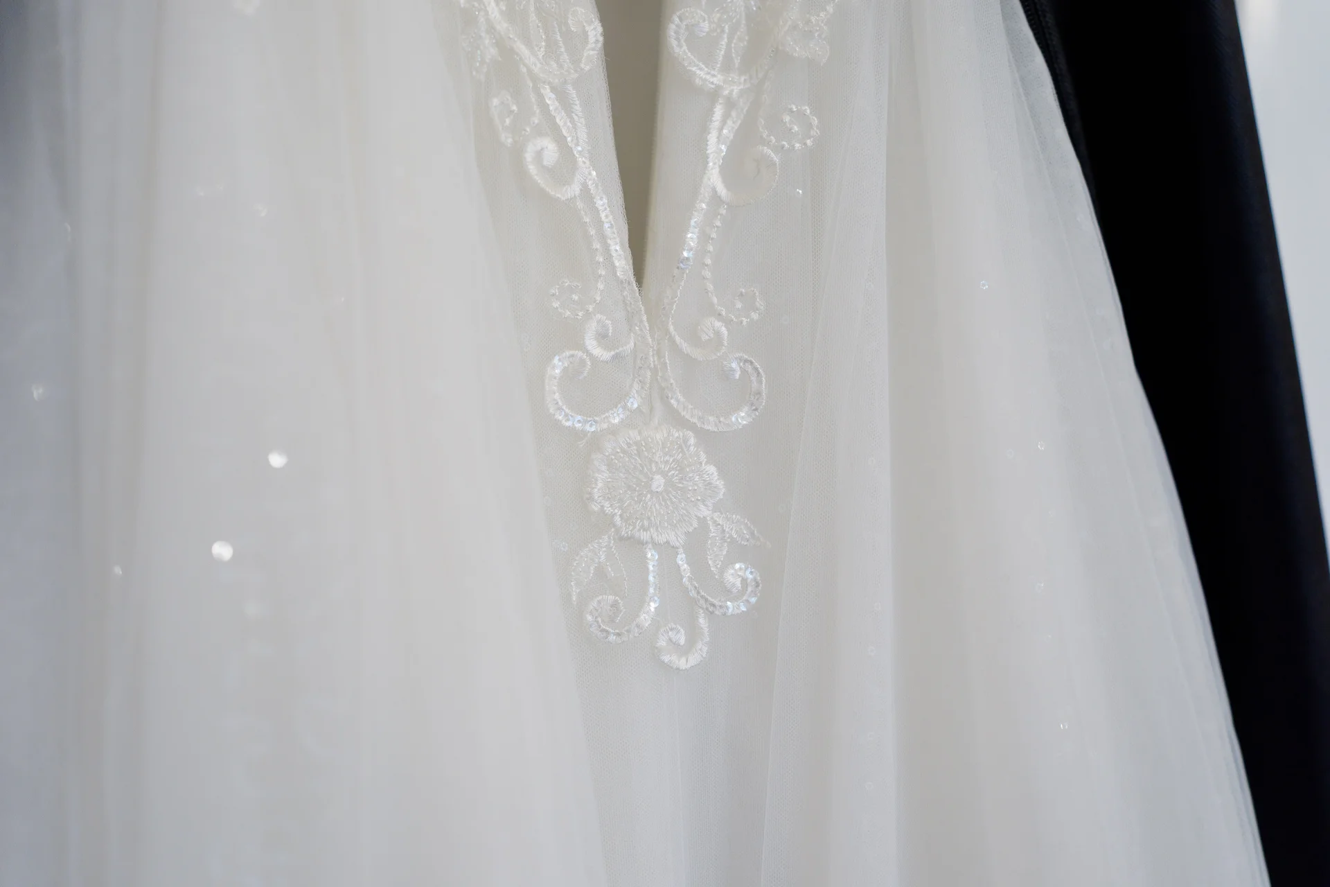 A close-up detail shot showcasing the delicate white embroidery and shimmering embellishments on a hanging wedding dress.