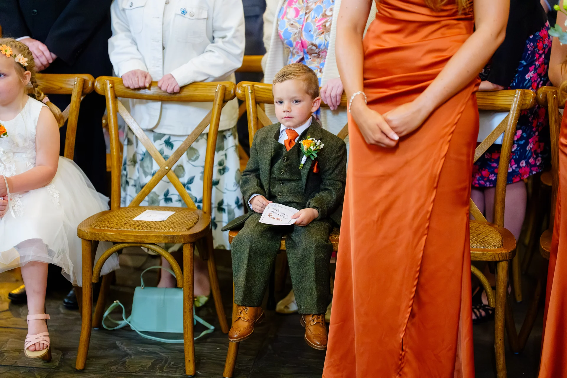 A young page boy in a smart green tweed suit with an orange tie sits on a wooden cross-back chair during a wedding ceremony.