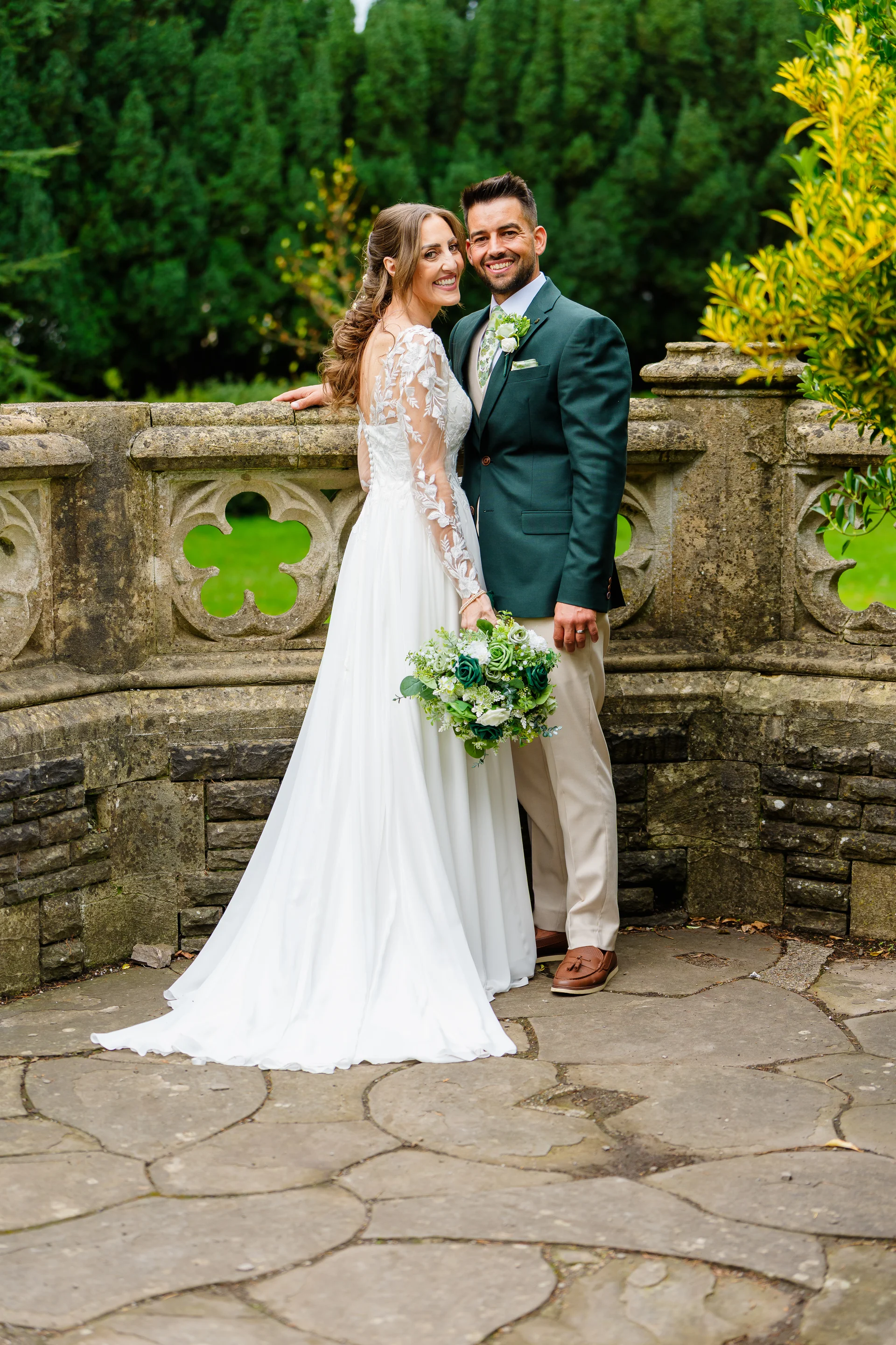 A happily smiling bride in a long-sleeved lace and tulle gown and a groom in a dark green suit jacket and beige trousers pose together on a stone patio against a lush garden backdrop.