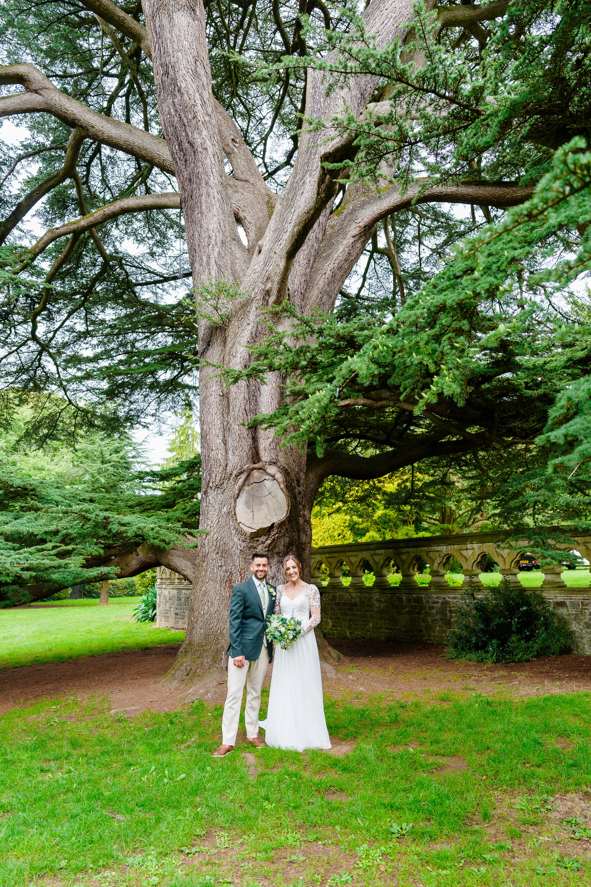 A smiling bride and groom stand hand in hand beneath the sprawling branches of an ancient cedar tree in a lush garden setting.