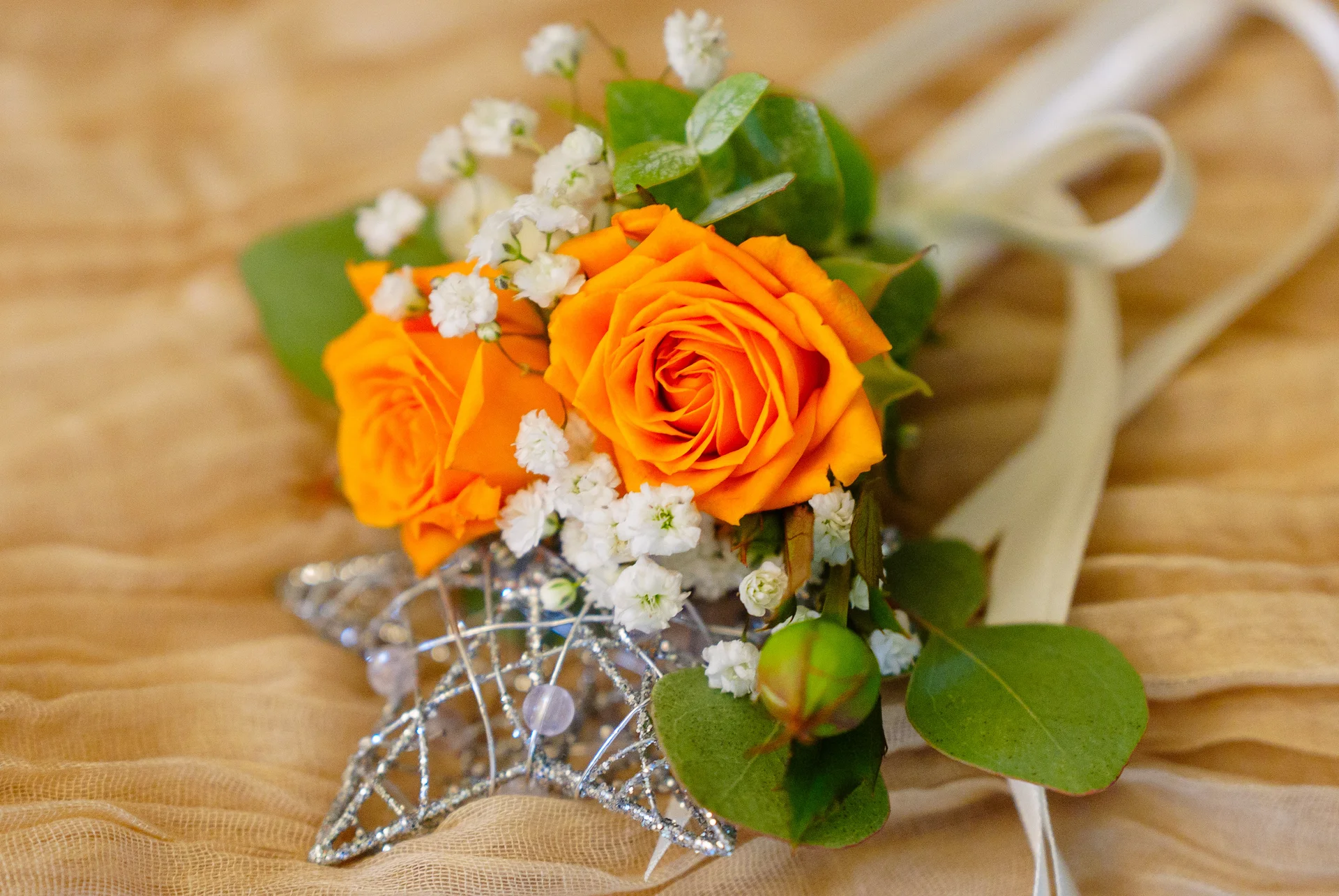 A close-up shot of a vibrant orange rose boutonniere accented with white baby's breath and green foliage, resting on a textured golden fabric surface.