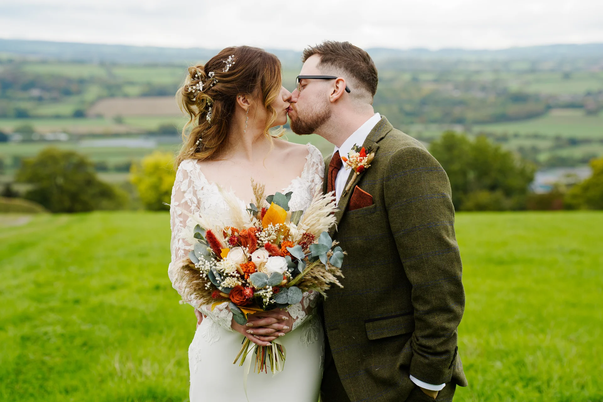 A newlywed couple shares a tender kiss outdoors against a scenic backdrop of rolling green hills during an autumn wedding portrait session.
