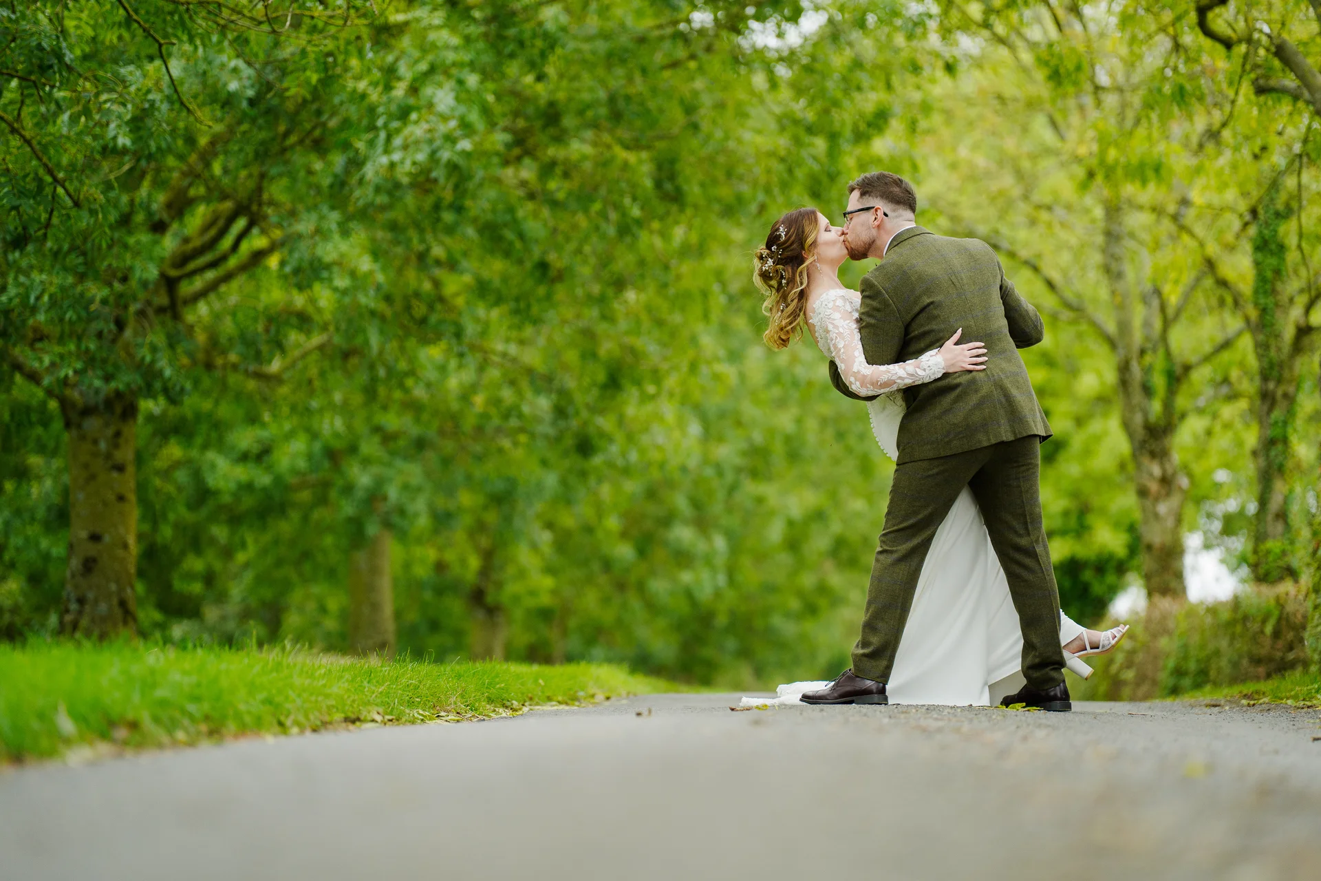 A newlywed couple shares a dramatic dip kiss along a tree-lined country path framed by lush green woodland.