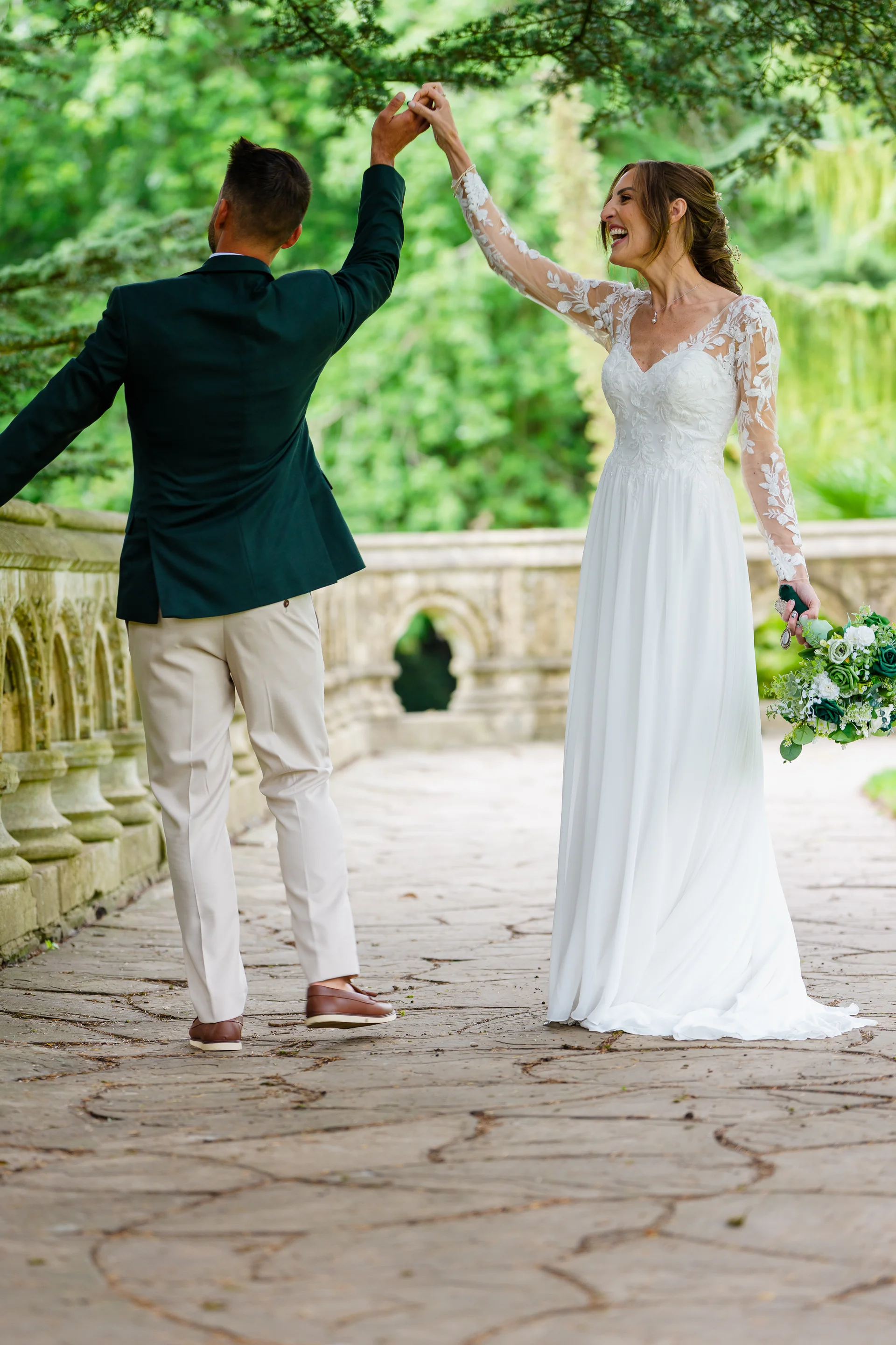 A newlywed couple shares a joyful dance outdoors on a stone bridge framed by lush greenery during their summer wedding portraits.