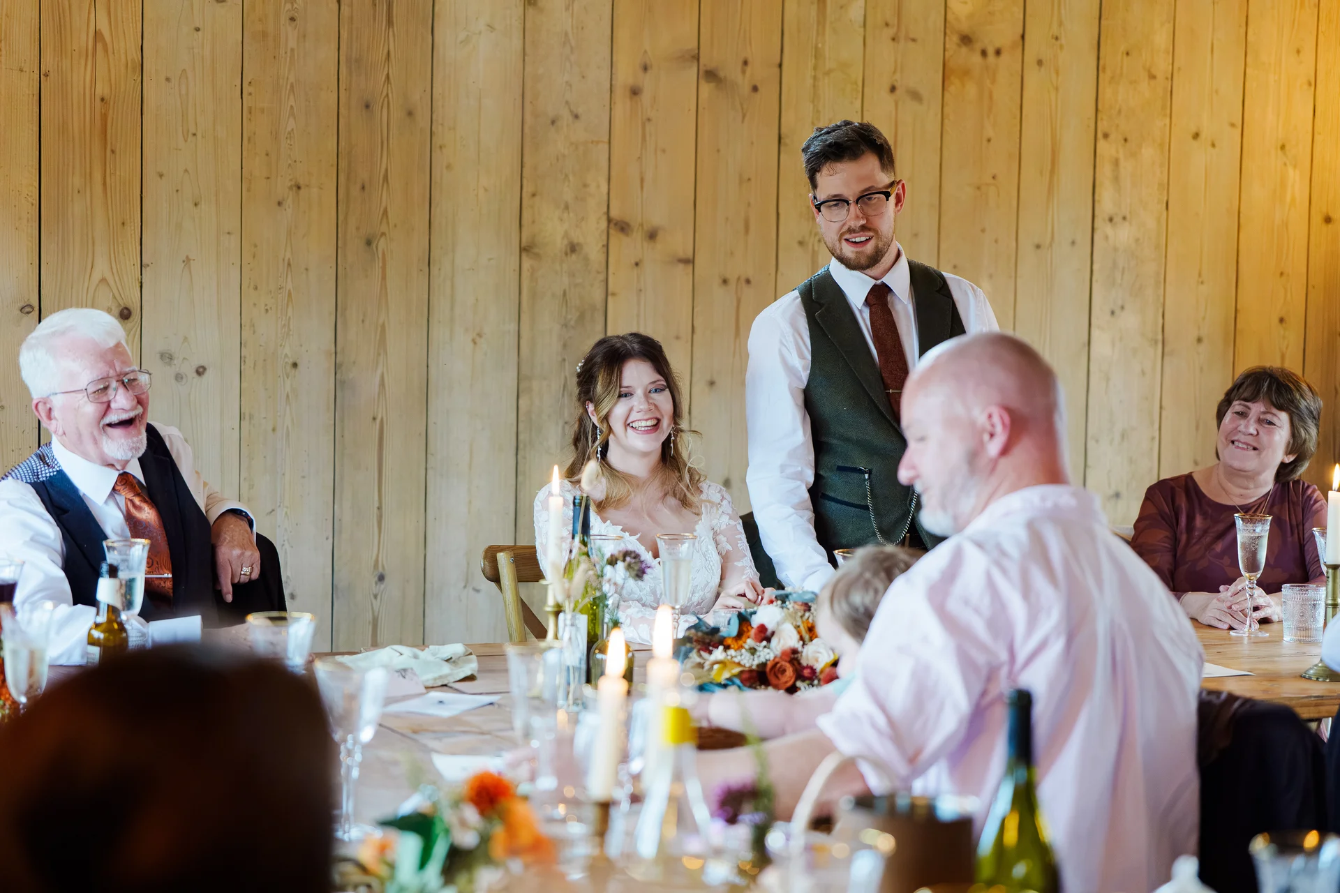 A groom stands to give a speech at the top table in a rustic wooden barn while the bride and family members laugh joyfully.