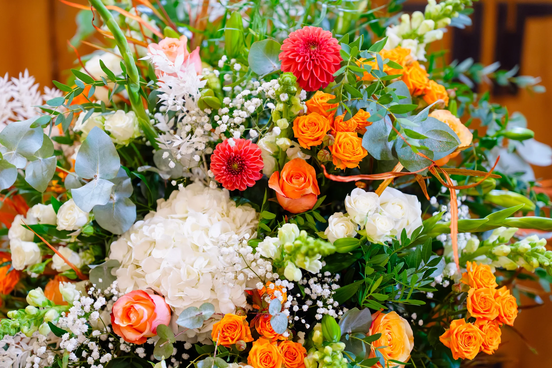 A vibrant wedding floral arrangement bursts with bright orange roses, deep red dahlias, white hydrangeas, and delicate baby's breath interspersed with lush eucalyptus leaves.