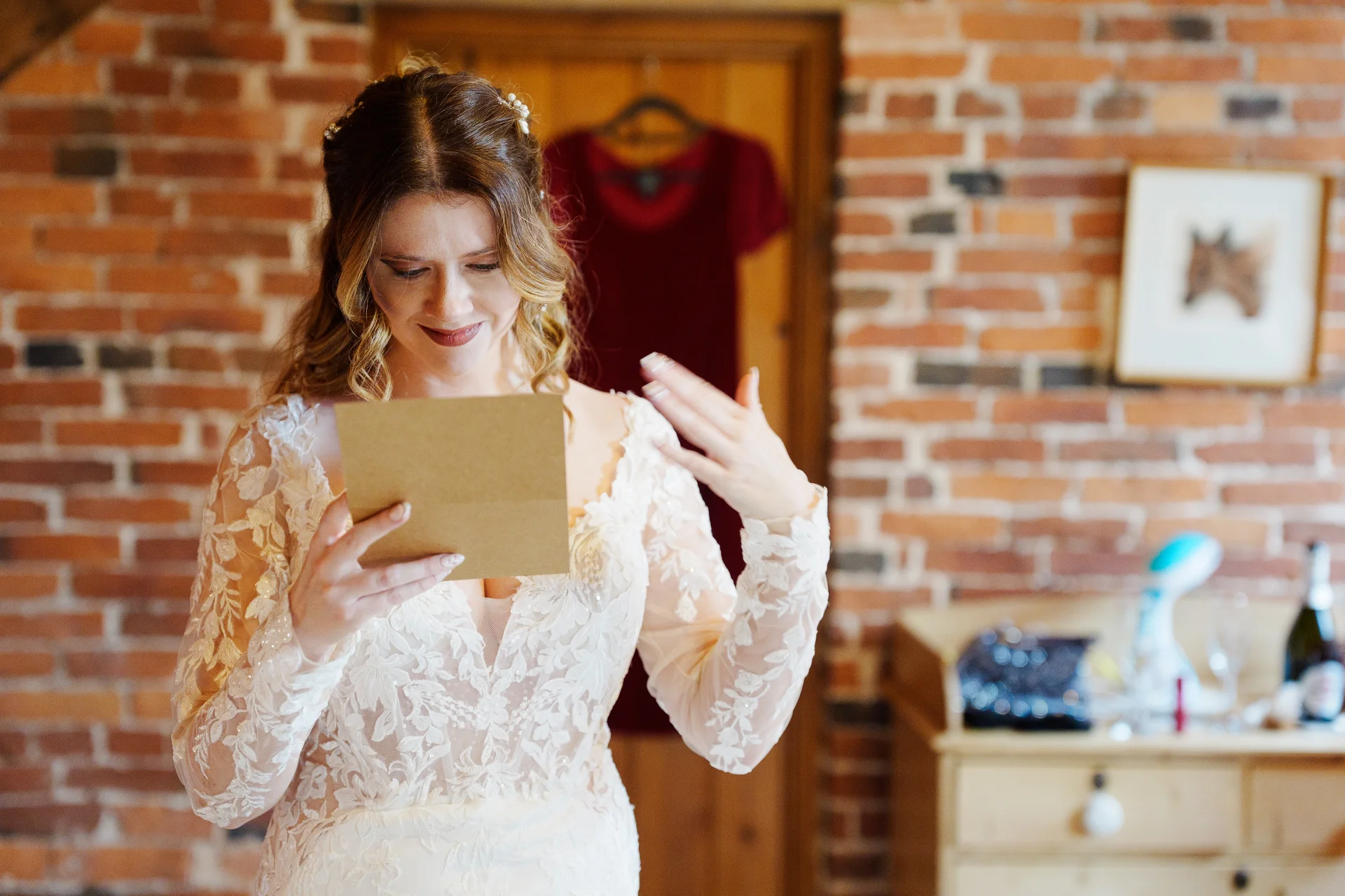 A smiling bride in a lace long-sleeve wedding dress reads a heartfelt letter while getting ready in an exposed brick room.
