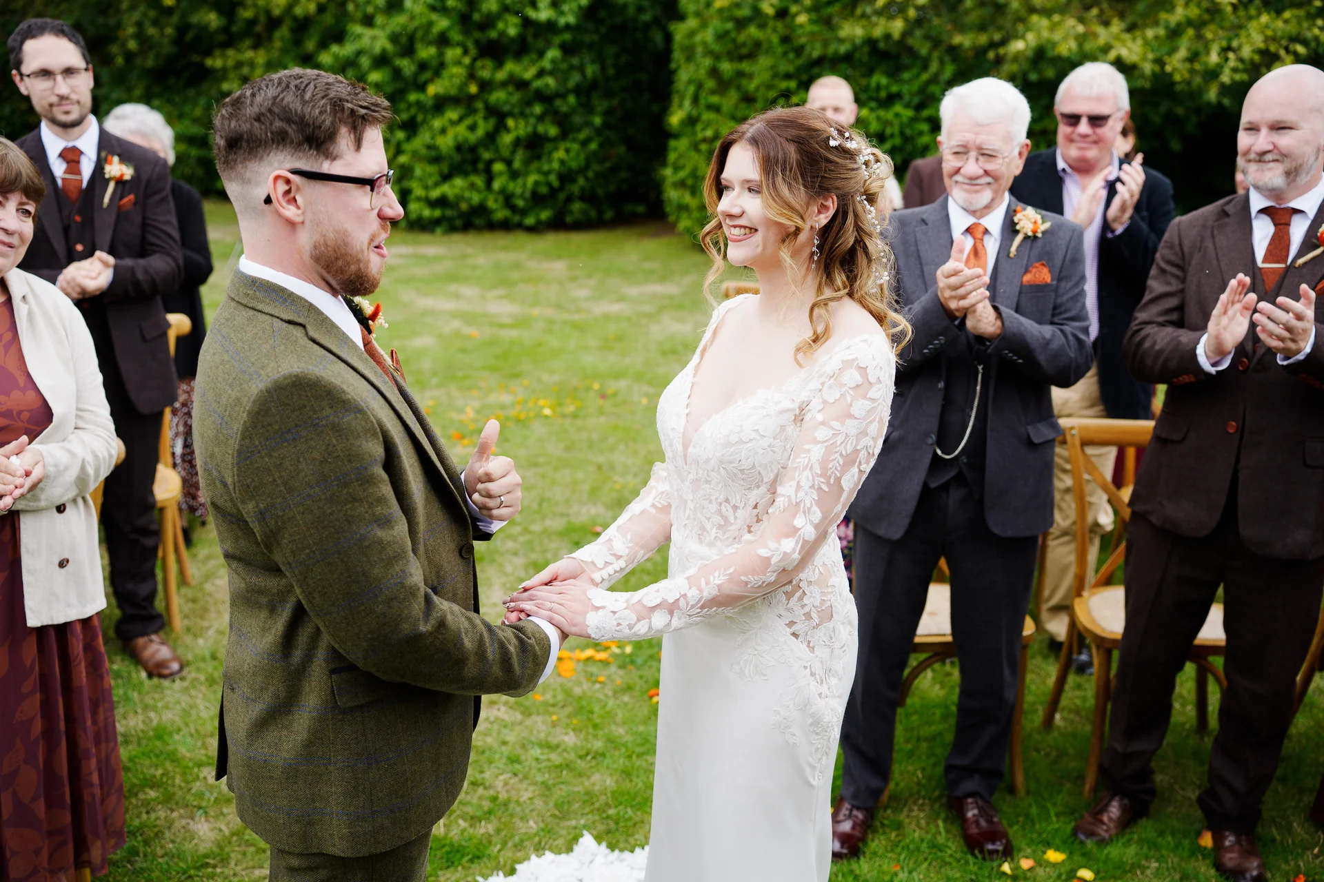 A smiling bride and groom hold hands during an outdoor garden ceremony while wedding guests clap and cheer in the background.