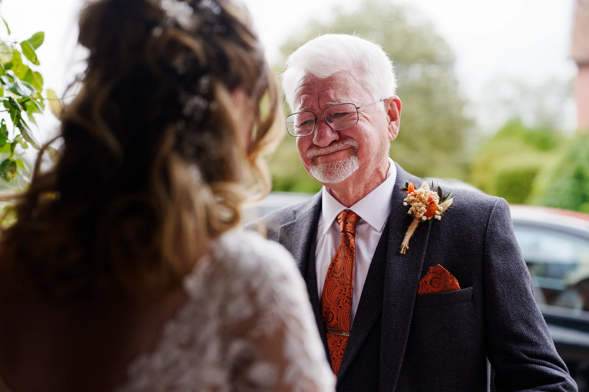 An emotional father of the bride wearing a grey tweed suit and patterned tie looks proudly at his daughter during a touching first look moment.