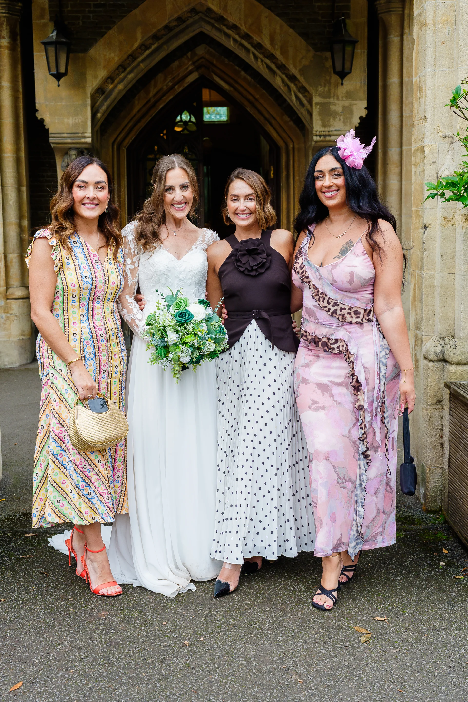 A smiling bride holding a green and white bouquet poses outside a stone church entrance alongside three bridesmaids wearing patterned and stylish dresses.