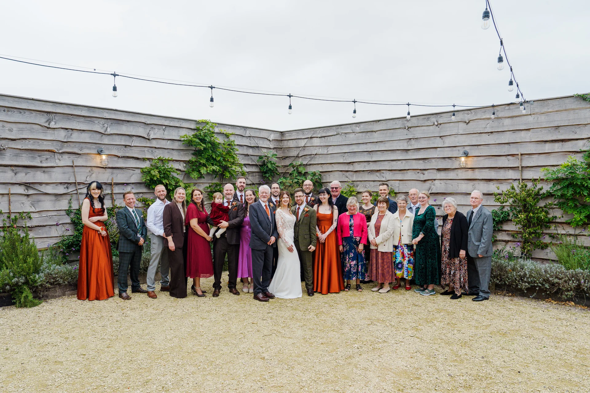 A large wedding group poses together outdoors on a gravel courtyard in front of a timber slat wall under string lighting.
