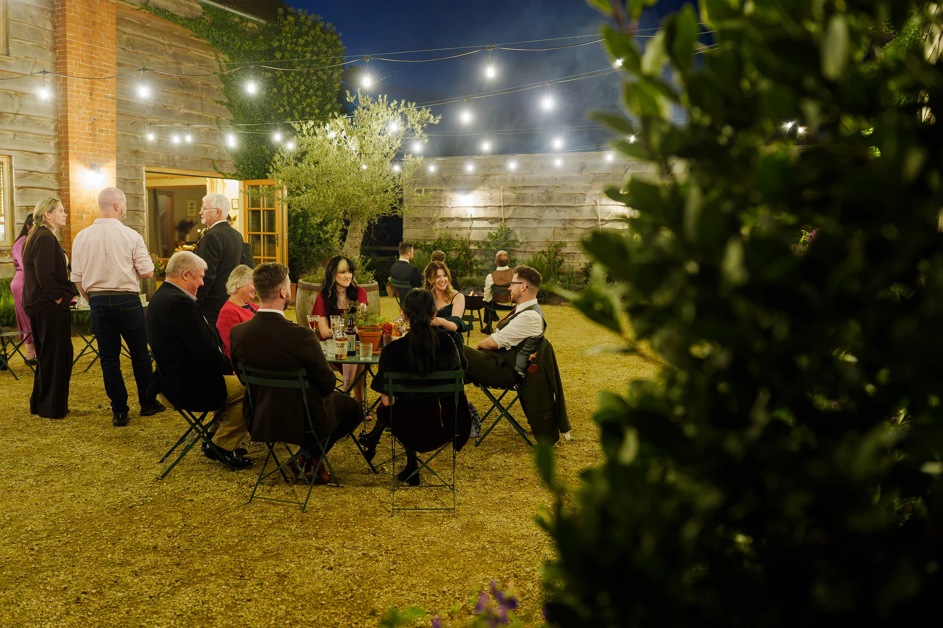 Wedding guests enjoy drinks and conversation in an outdoor courtyard lit by overhead festoon lights during an evening reception.