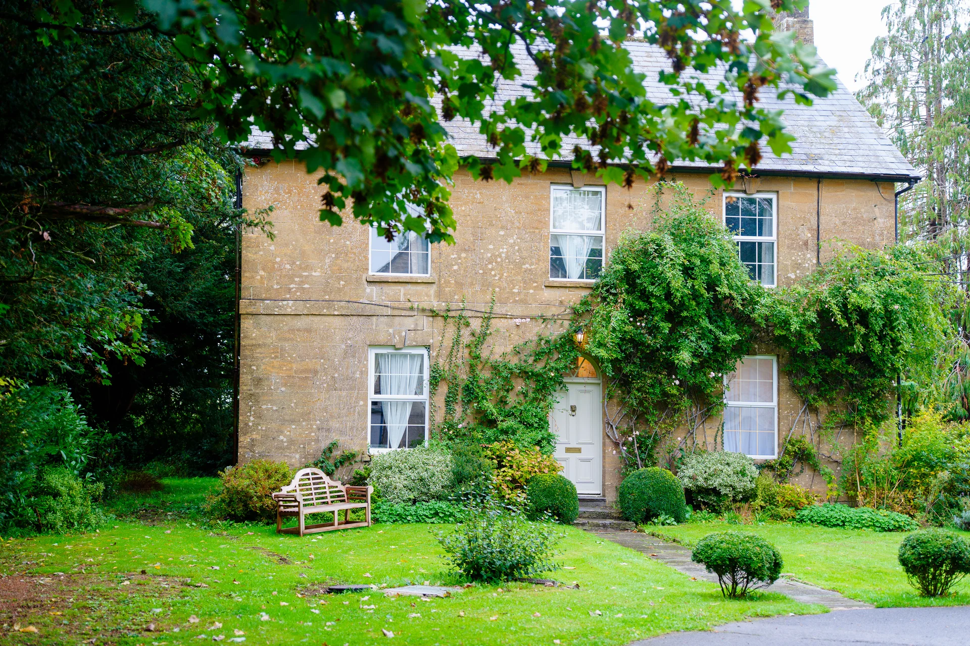 A charming stone country house with climbing green foliage surrounding the front door and a wooden bench on the manicured lawn.